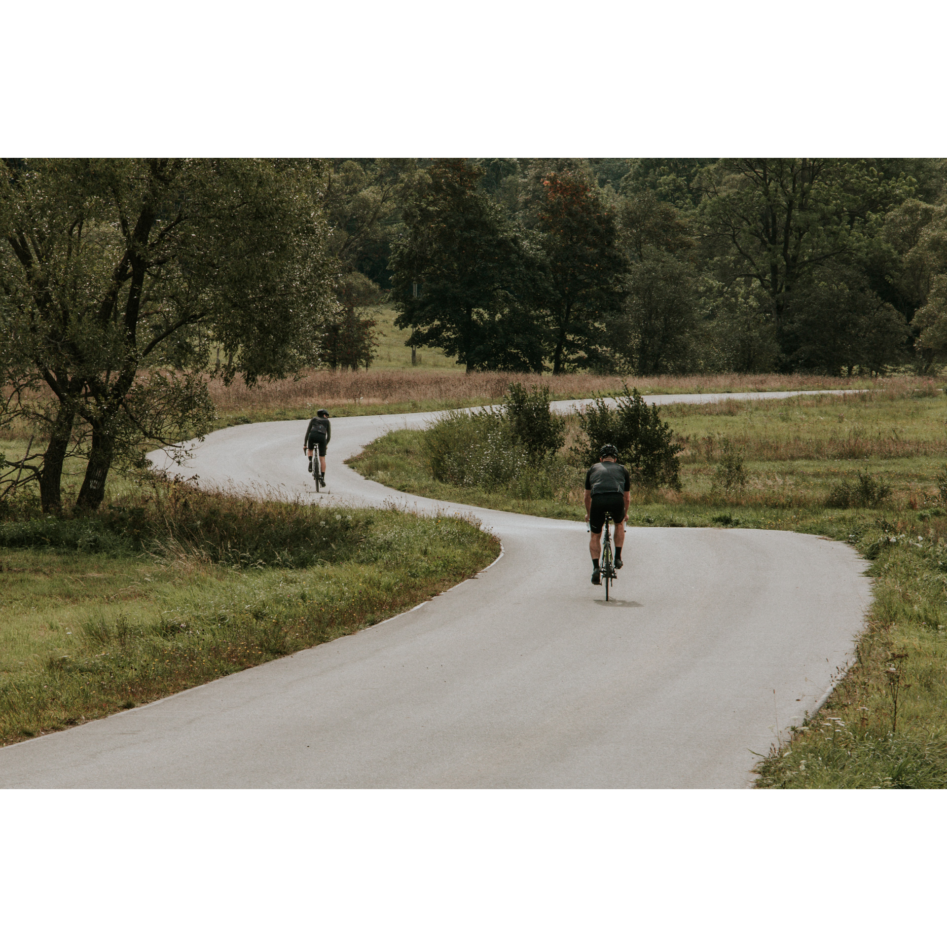 Two cyclists in black clothes and helmets riding bicycles along an asphalt winding road among green meadows, trees and hills