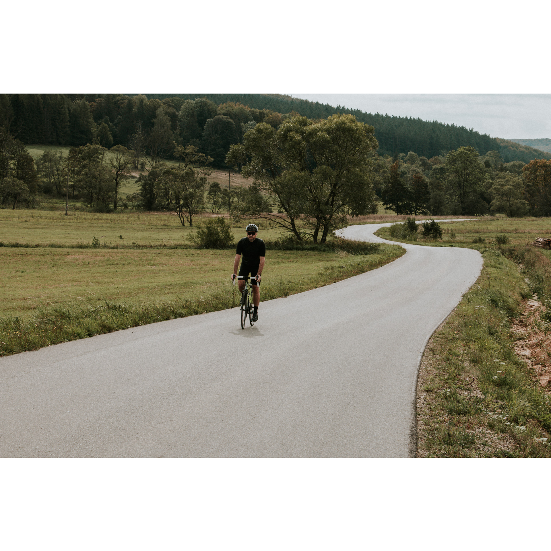 Kolarz w czarnym stroju i kasku jadący na rowerze asfaltową krętą drogą wśród zielonych łąk, drzew i wzgórz