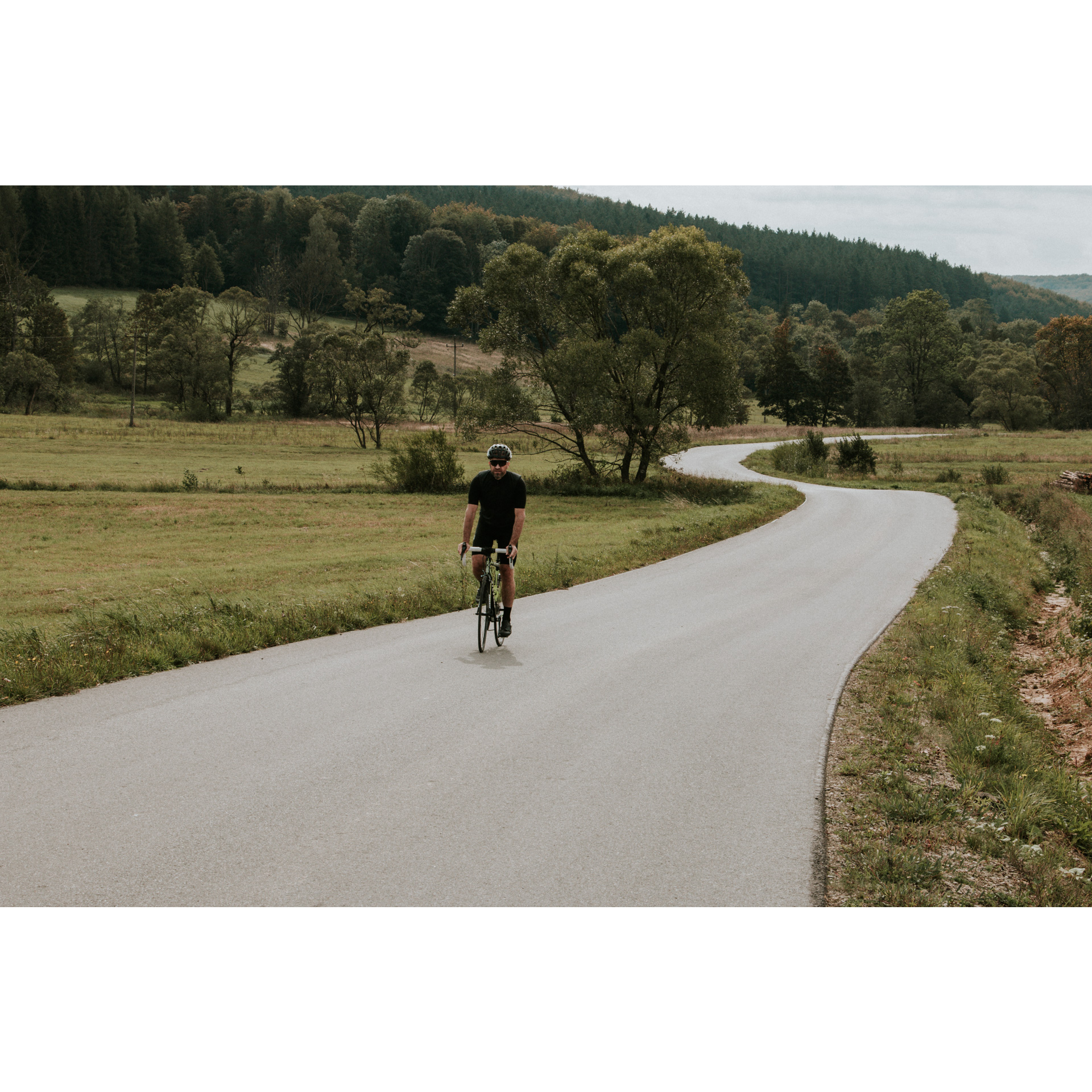 A cyclist in a black outfit and helmet cycling along an asphalt winding road among green meadows, trees and hills