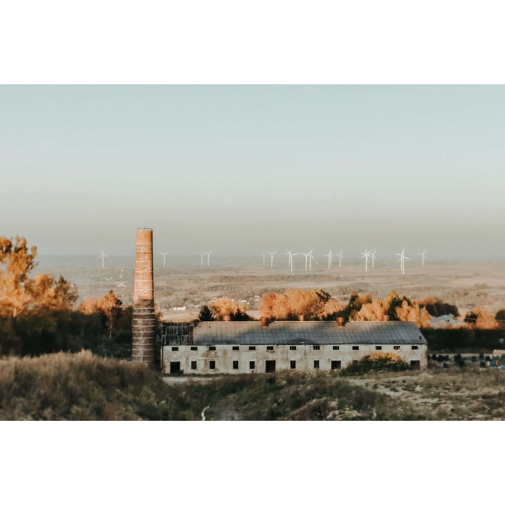 A bright, oblong building with many windows and a tall chimney on the left, hills and a field with many wind turbines in the background