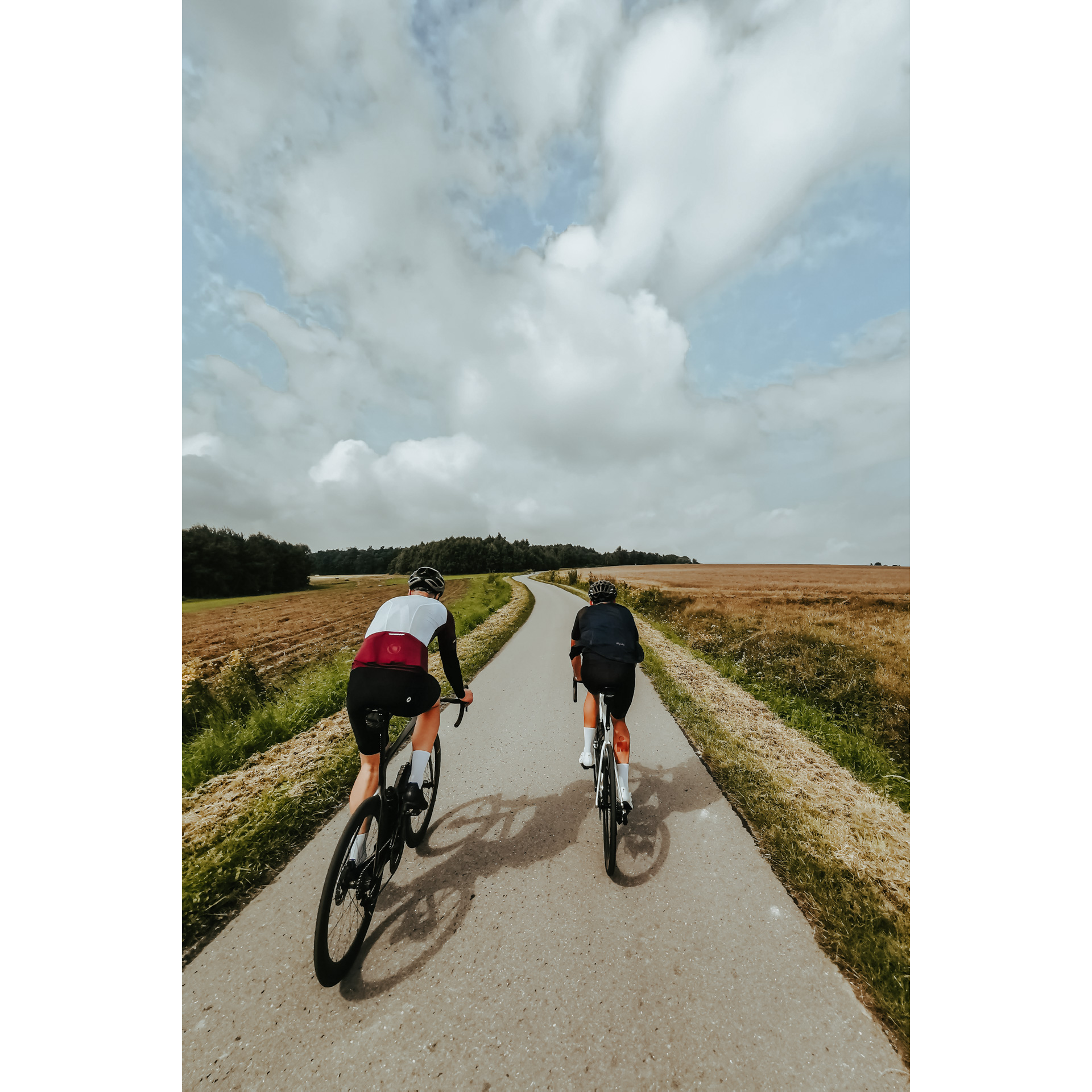 Two cyclists in cycling clothes and helmets riding side by side on a narrow asphalt road among agricultural fields towards the forest