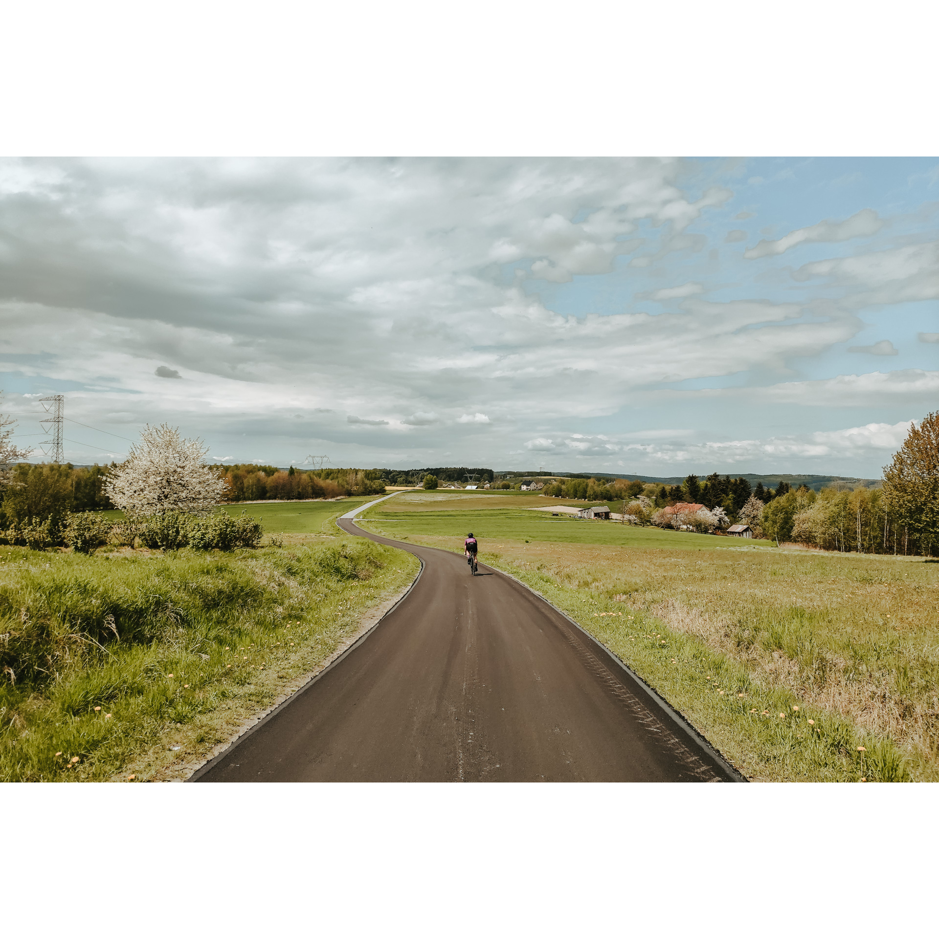 A cyclist in a black outfit and helmet cycling along an asphalt road running between green meadows, blue sky and clouds in the background, houses and hills in the distance