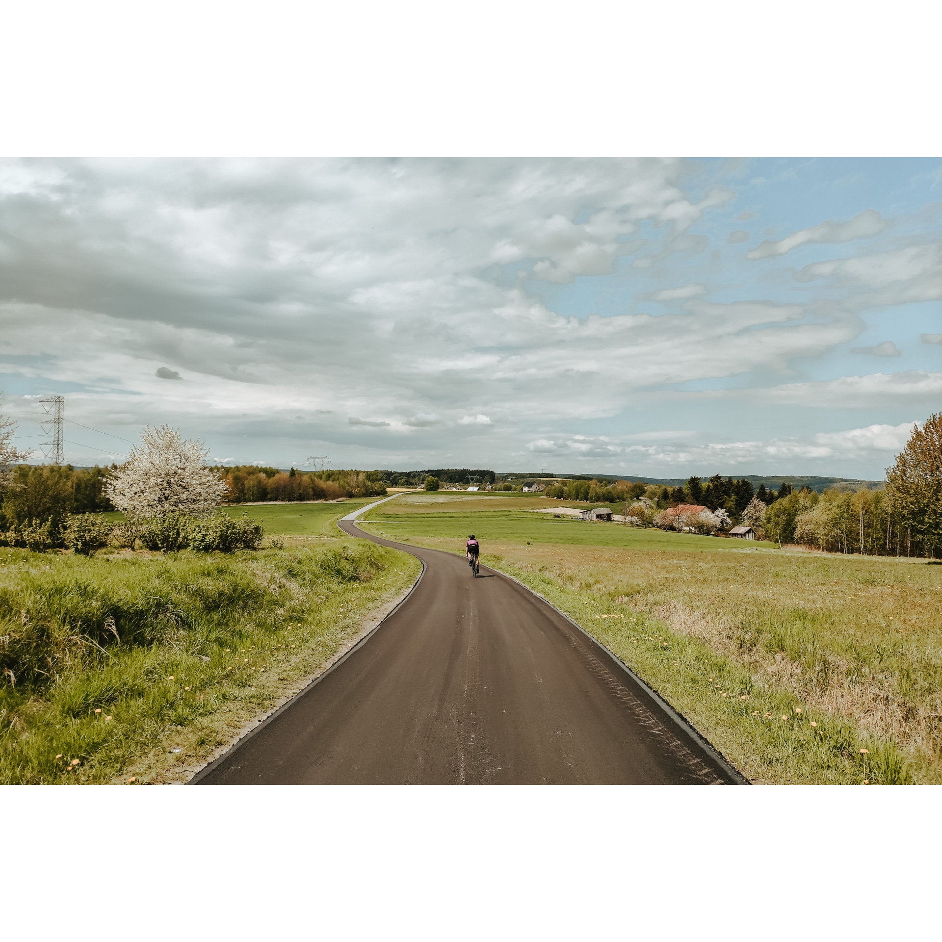 Rowerzysta w czarnym stroju i kasku jadący na rowerze drogą asfaltową biegnącą między zielonymi łąkami, w tle niebieskie niebo i chmury, w oddali domostwa i wzgórza