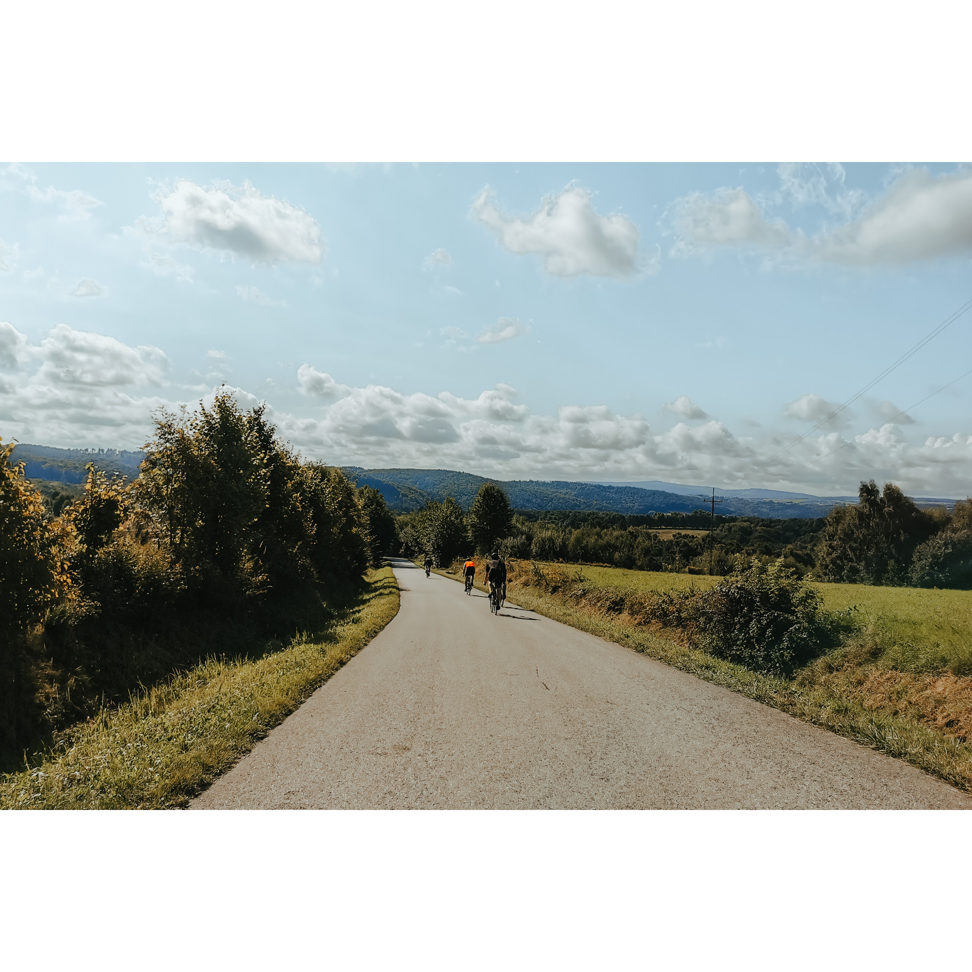5 rowerzystów w strojach kolarskich jadących z górki drogą asfaltową wśród zielonych łąk i drzew, w tle wzgórza i błękitne niebo oraz chmury