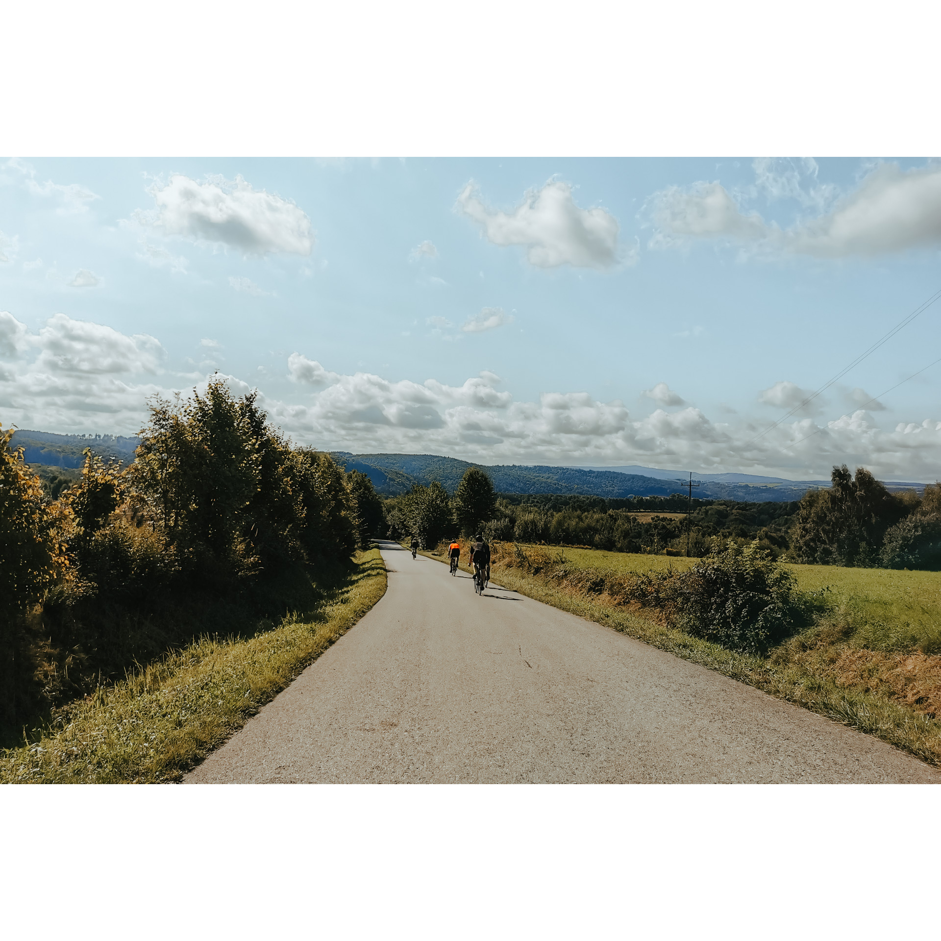 5 cyclists in cycling clothes going downhill on an asphalt road among green meadows and trees, in the background hills and blue sky and clouds