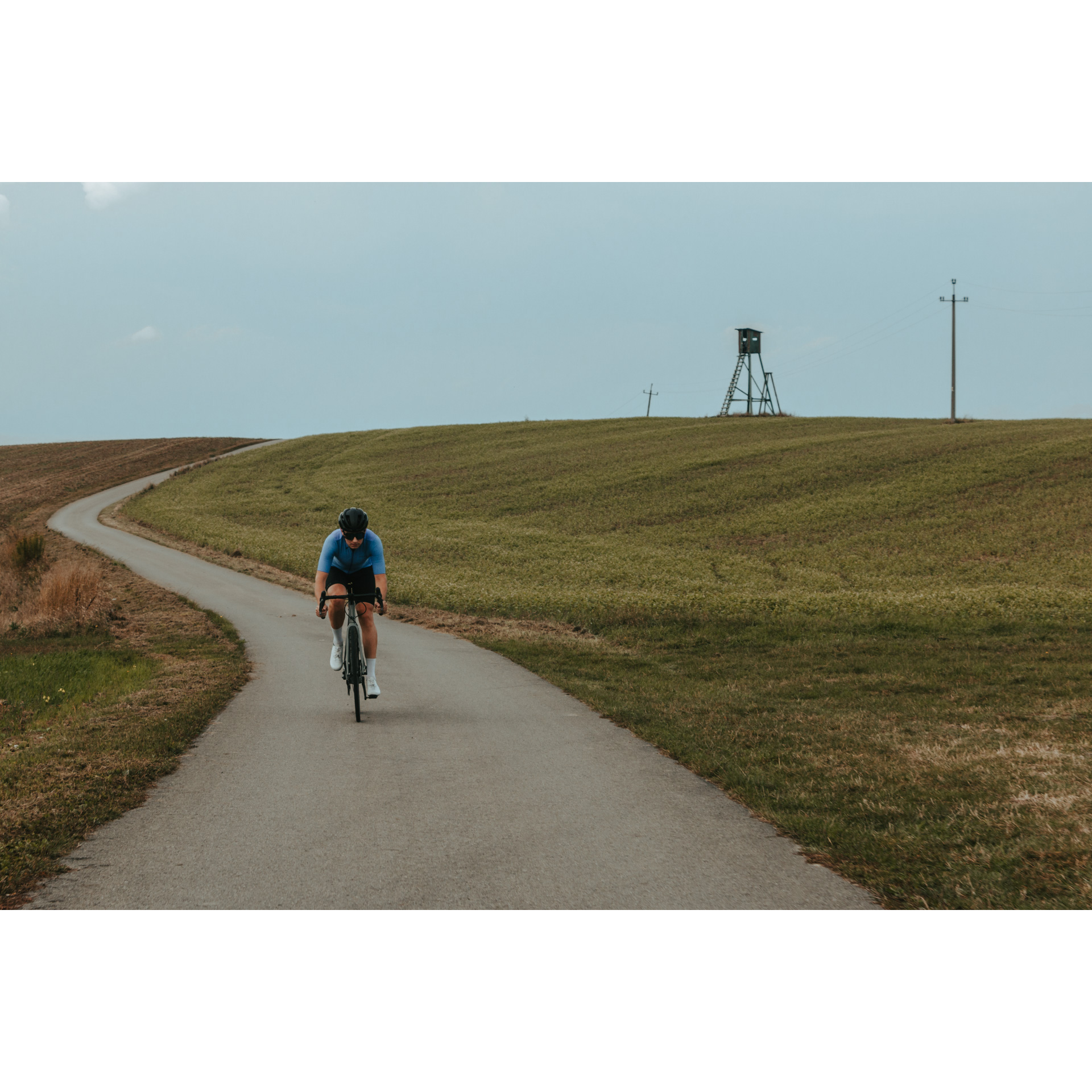 Rowerzysta w niebiesko-czarnym stroju kolarskim, kasku i okularach jadący rowerem asfaltową drogą wśród zielonych pagórków, w oddali na pagórku ambona obserwacyjna