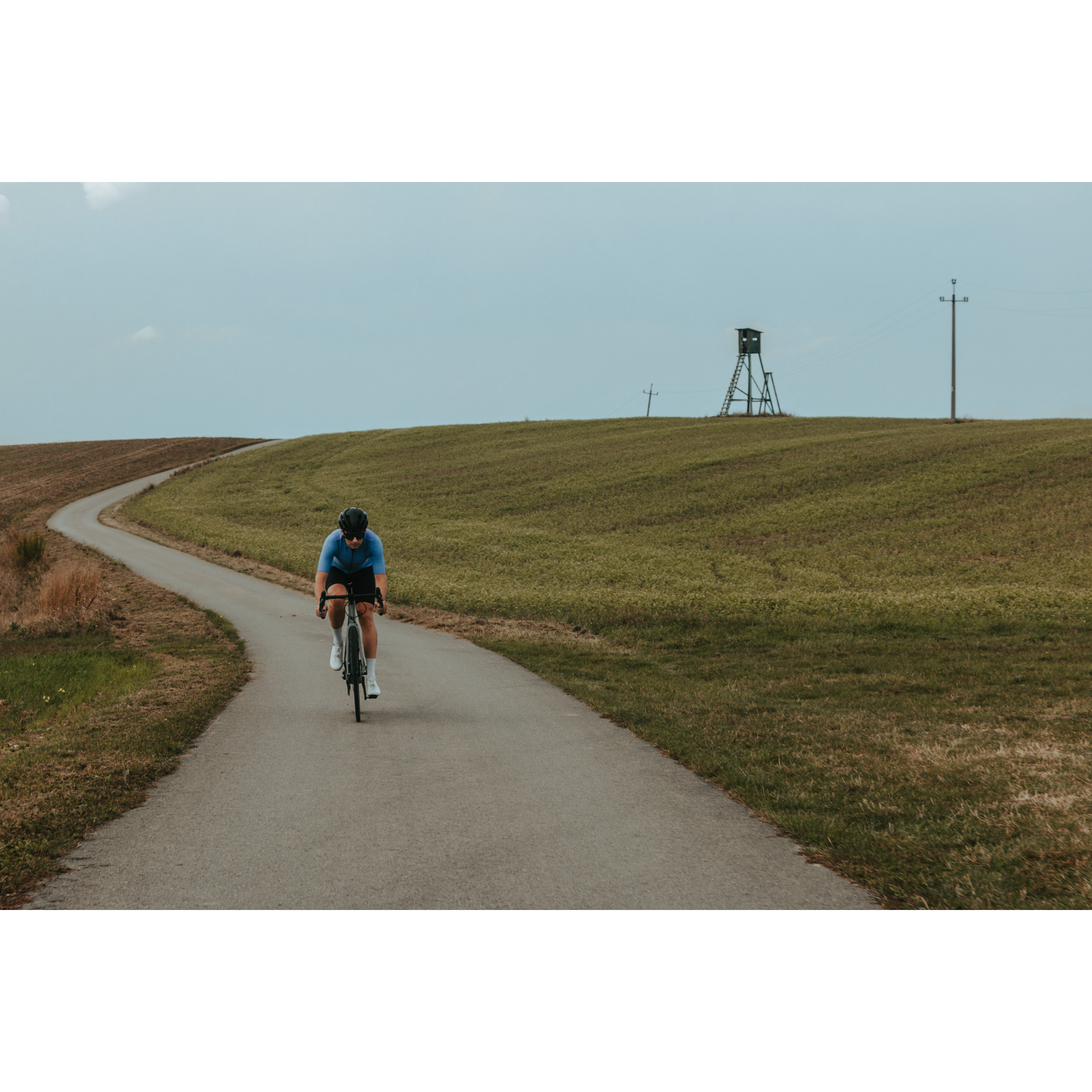 A cyclist in a blue and black cycling outfit, helmet and glasses riding a bicycle on an asphalt road among green hills, in the distance there is an observation tower on a hill