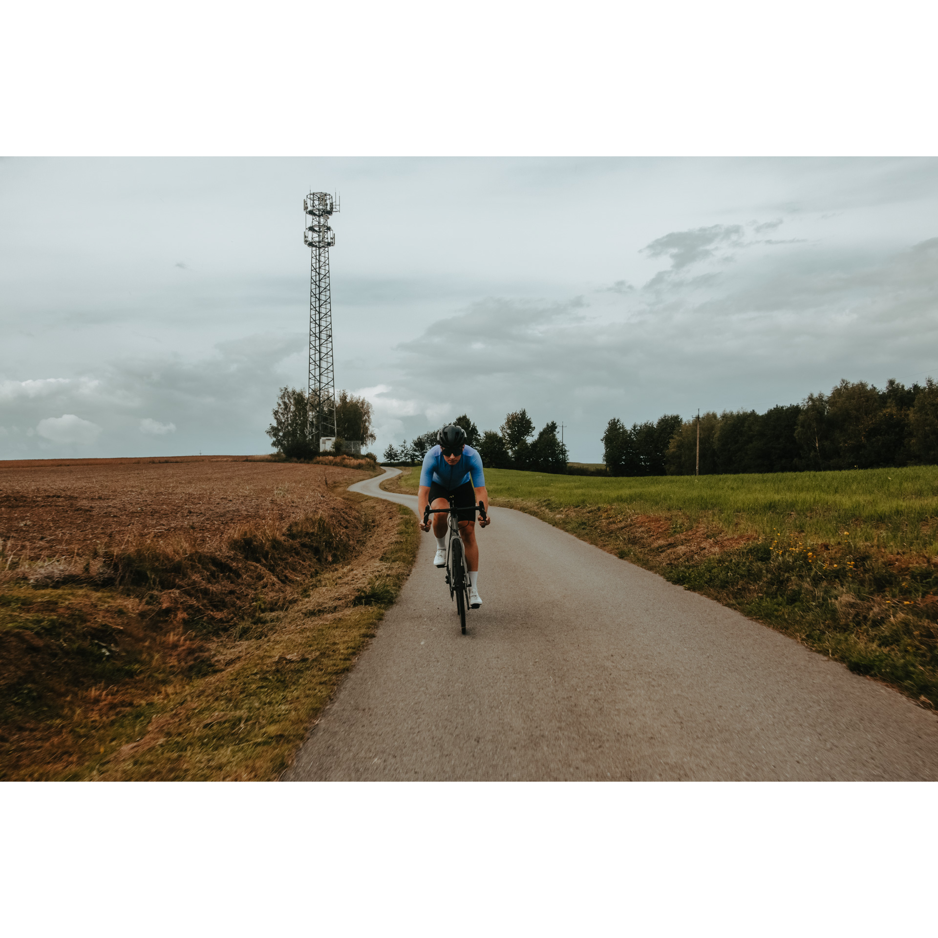 Rowerzysta w niebiesko-czarnym stroju kolarskim kasku i okularach jadący rowerem asfaltową drogą wśród łąk i pól, w tle drzewa, szare niebo i wysoki słup elektryczny