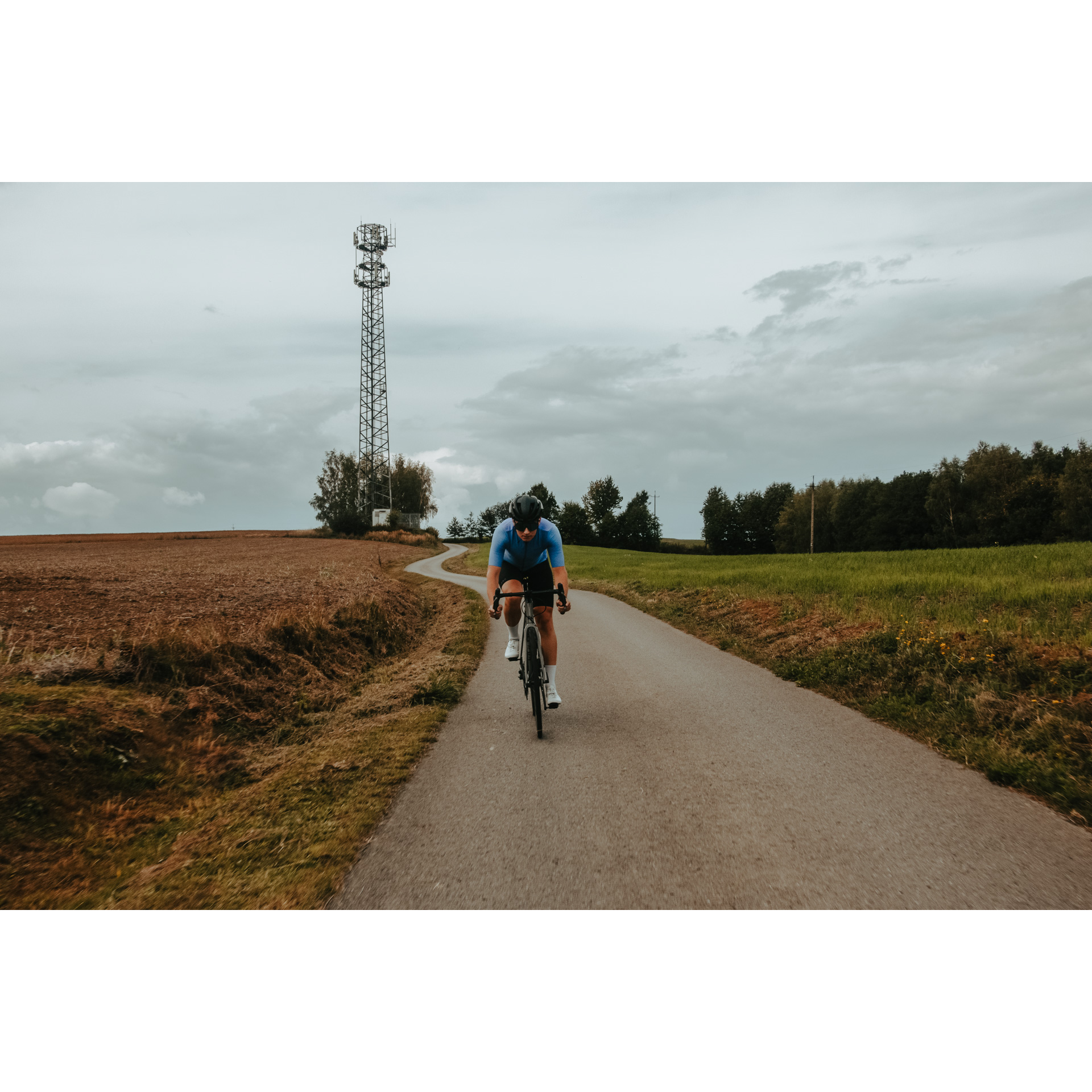 A cyclist in a blue and black cycling outfit, helmet and glasses riding a bicycle on an asphalt road among meadows and fields, in the background trees, gray sky and a high electric pole