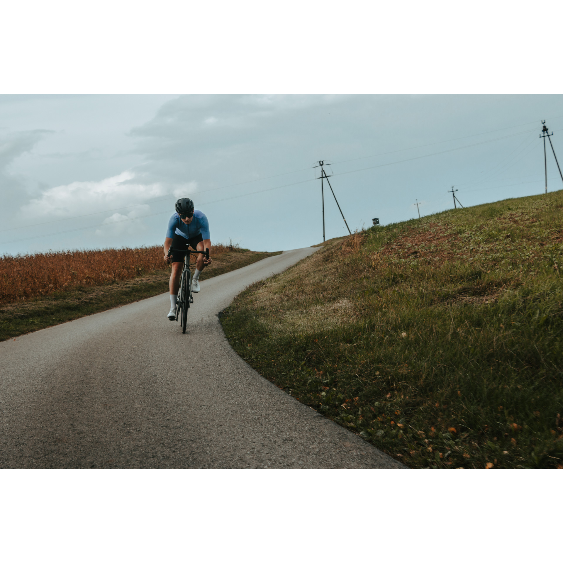 Rowerzysta w niebiesko-czarnym stroju kolarskim, kasku i okularach zjeżdżający z górki rowerem asfaltową drogą wśród zielonych łąk
