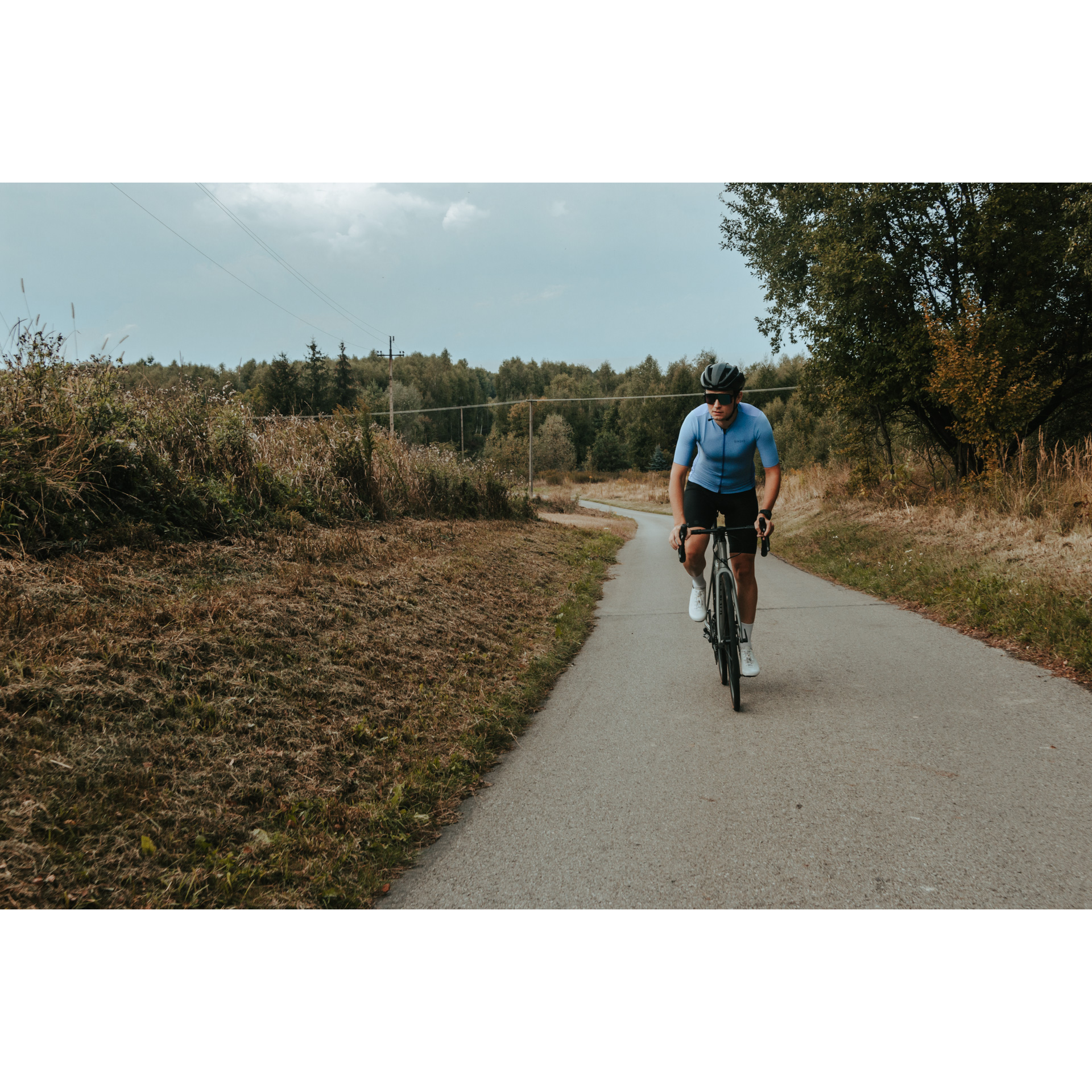 Rowerzysta w niebiesko-czarnym stroju kolarskim, kasku i okularach jadący rowerem asfaltową od strony lasu wśród krzewów i drzew