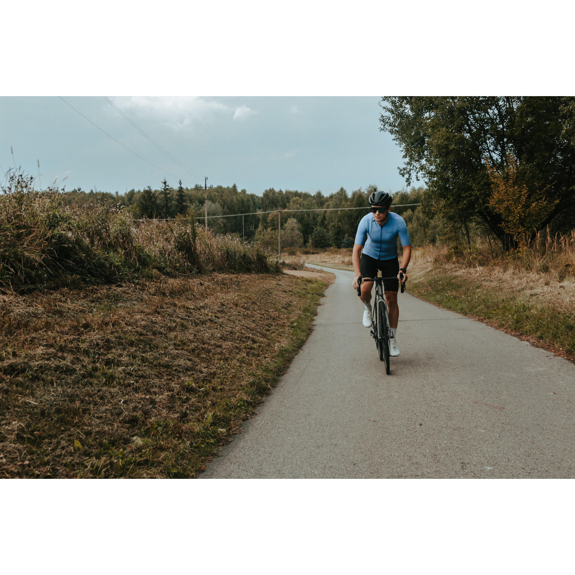 A cyclist in a blue and black cycling outfit, helmet and glasses riding a bicycle on an asphalt road from the forest among bushes and trees