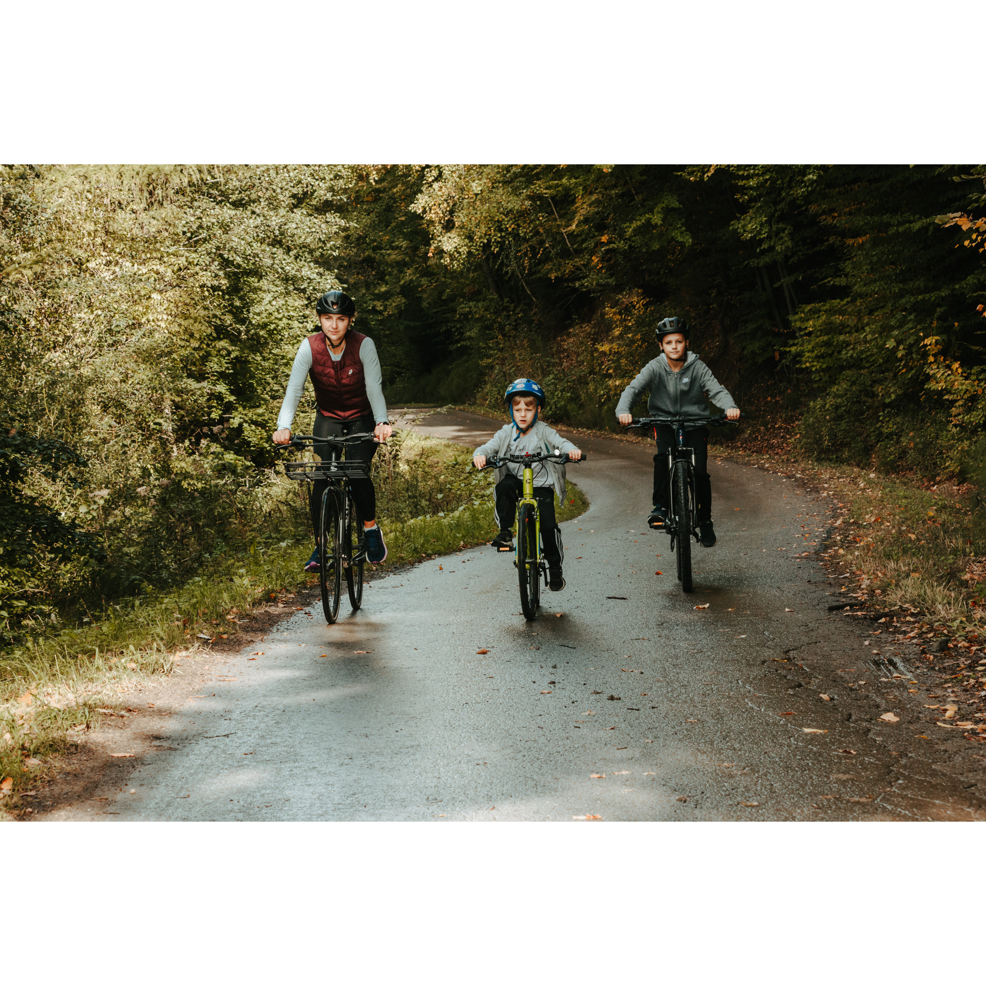 A woman and two boys in helmets cycling along an asphalt, wet road among green trees