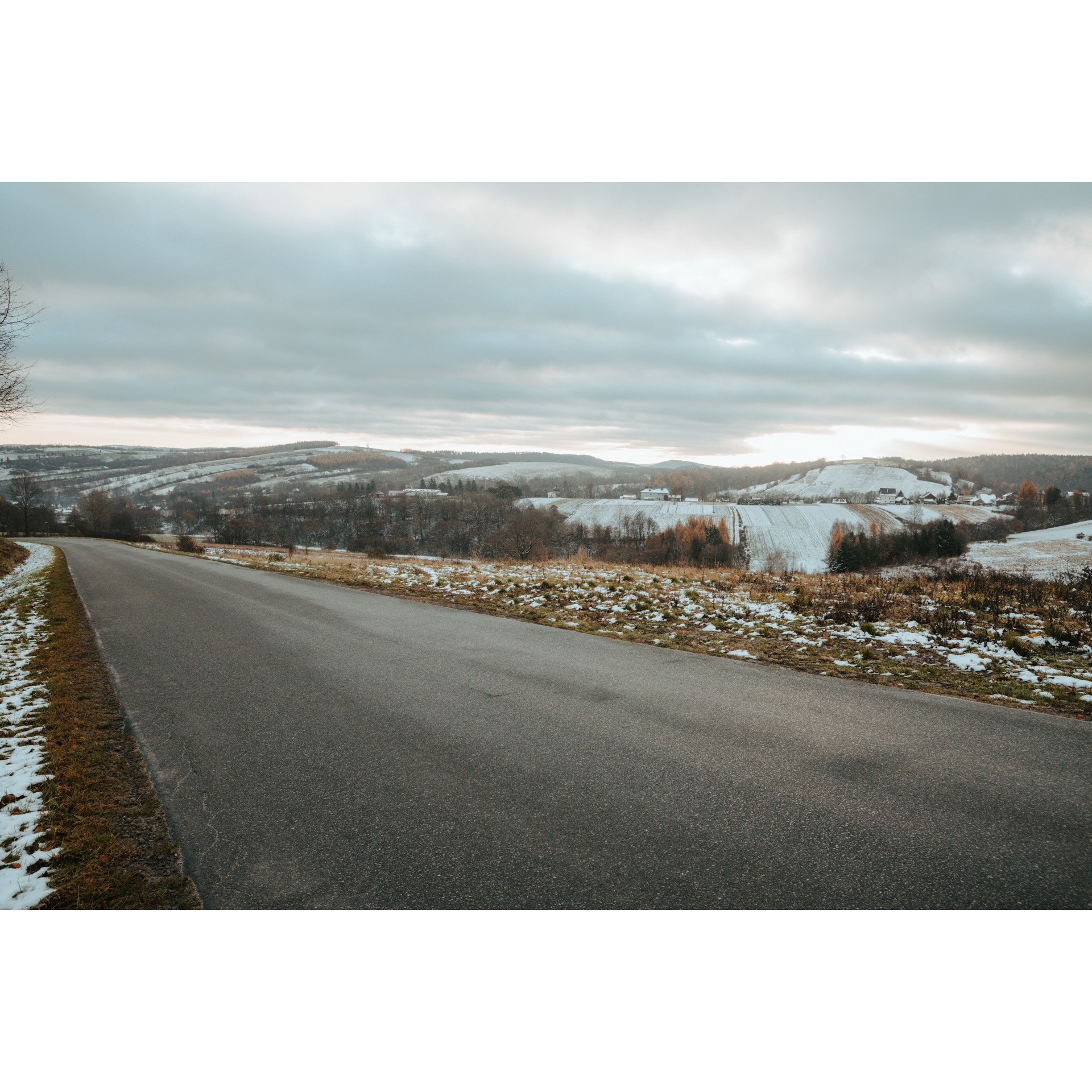 An asphalt road with a view of lightly snowy hills and trees without leaves and a cloudy sky