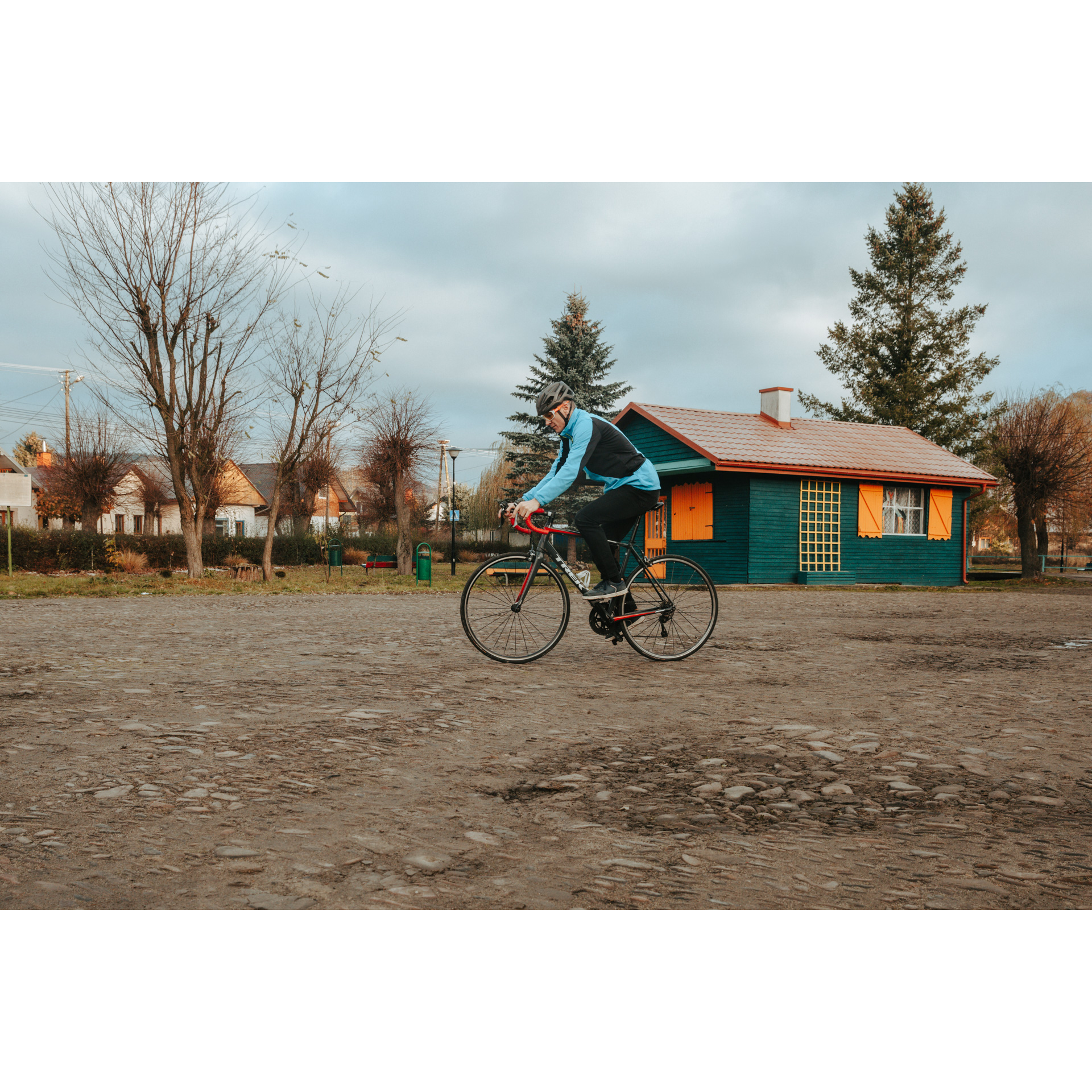 Rowerzysta w niebiesko-czarnej kurtce, kasku i okularach jadący po piaszczystym placu, w tle zielony, drewniany budynek z pomarańczowymi okiennicami, w oddali inne drewniane budynki
