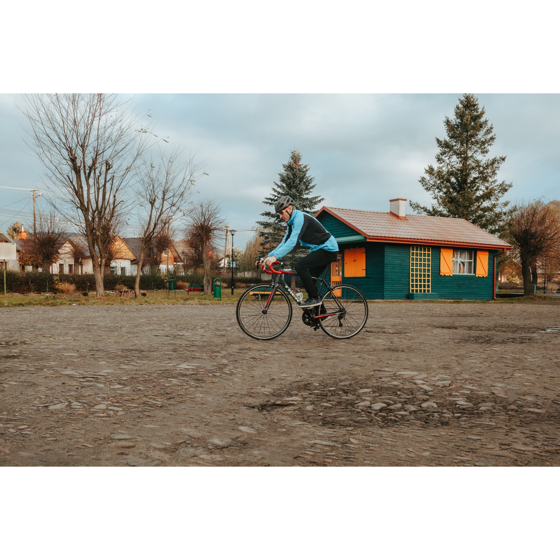 A cyclist in a blue and black jacket, helmet and glasses riding on a sandy square, in the background a green wooden building with orange shutters, in the distance other wooden buildings