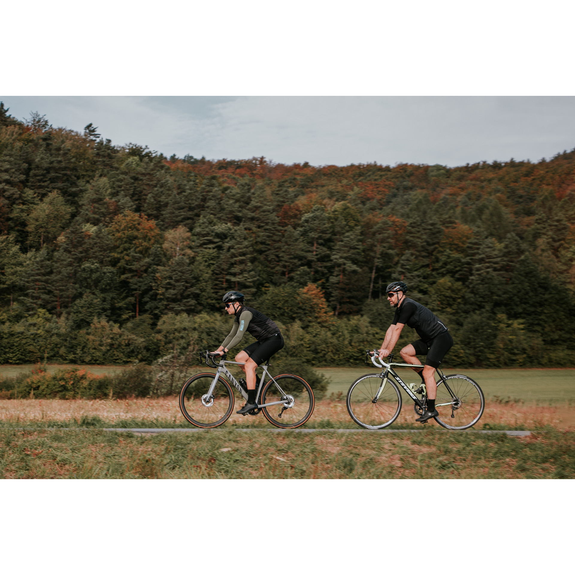 Dwóch rowerzystów w czarnych strojach kolarskich i kaskach jadących po asfaltowej drodze biegnącej między zielonymi łąkami, w tle drzewa na wzgórzu