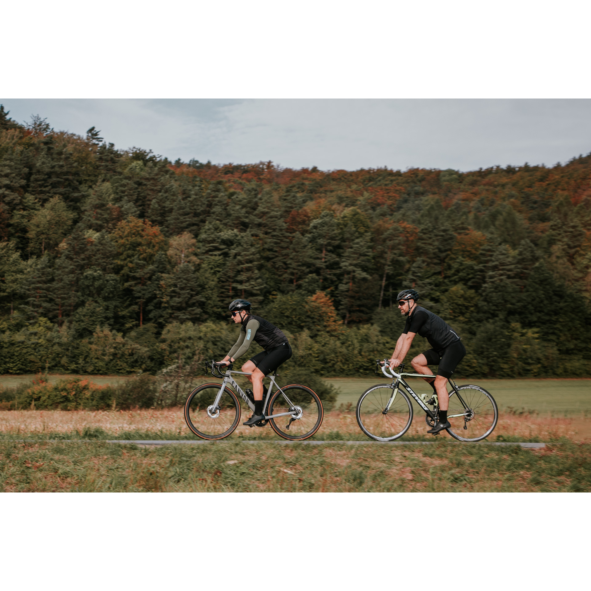 Two cyclists in black cycling clothes and helmets riding on an asphalt road running between green meadows, trees on a hill in the background