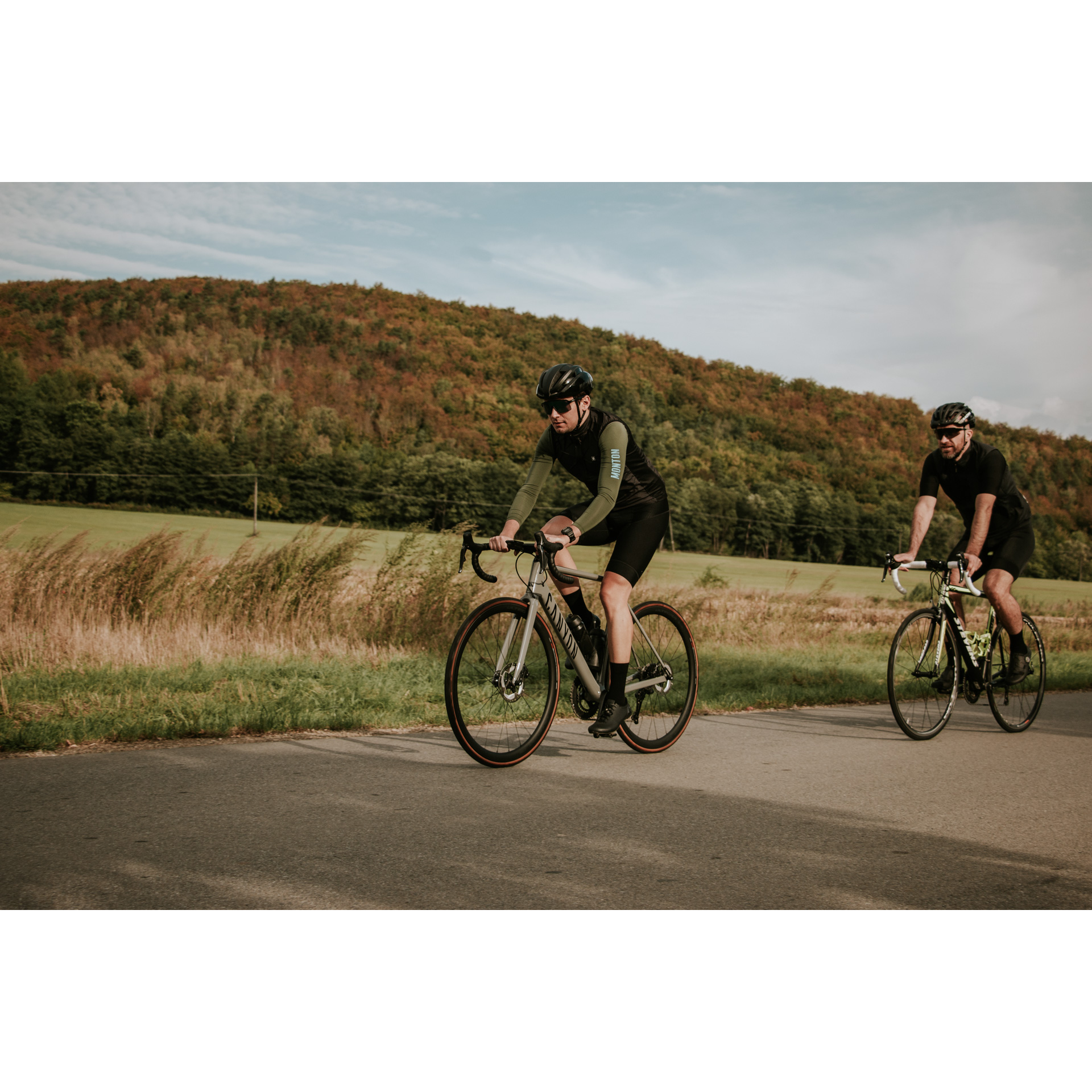 Dwóch rowerzystów w czarnych strojach kolarskich i kaskach jadących po asfaltowej drodze biegnącej wzdłuż łąk, w tle wzgórze porośnięte brązowo-zielonymi drzewami