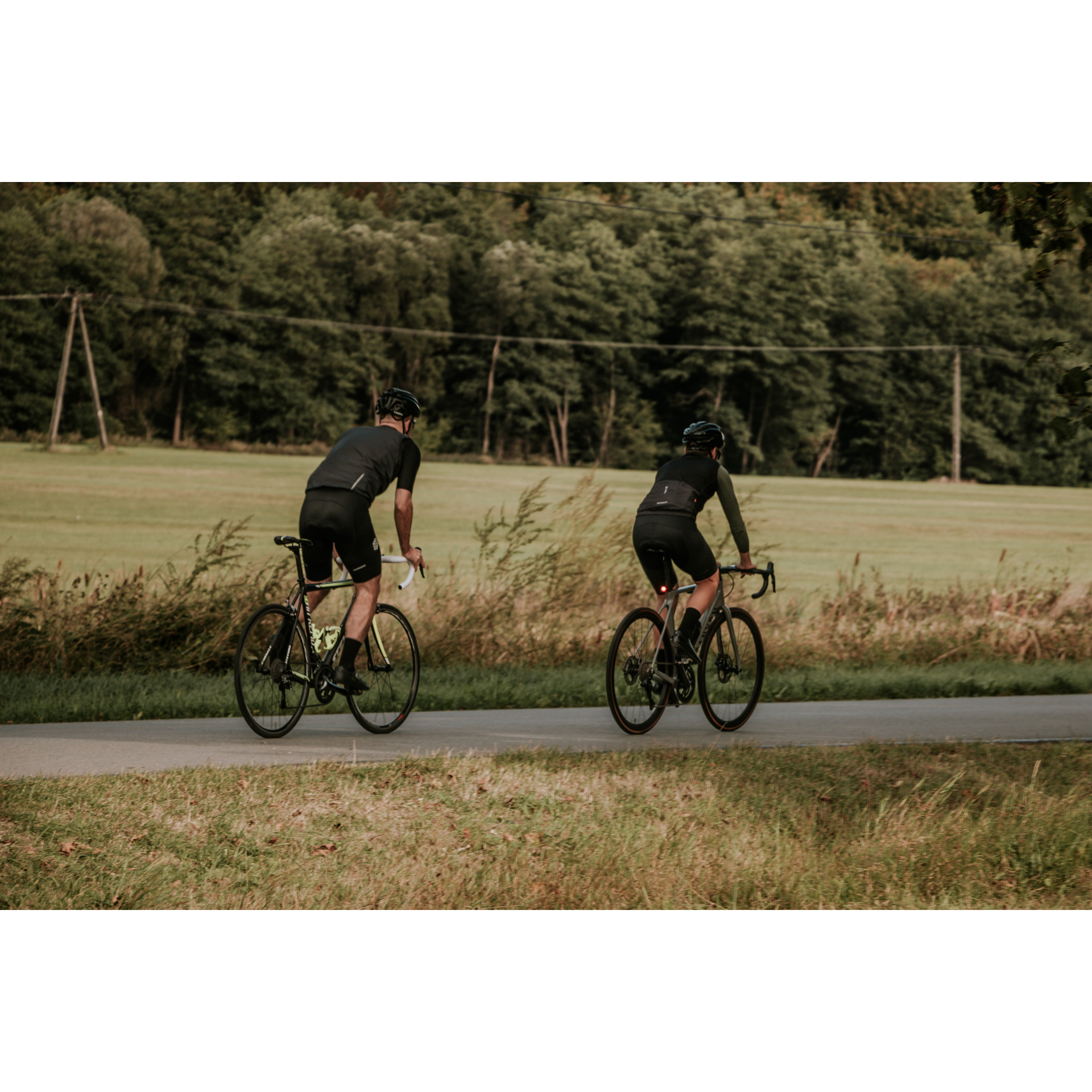 Two cyclists in black cycling clothes and helmets riding on an asphalt road running between green meadows, green trees in the background