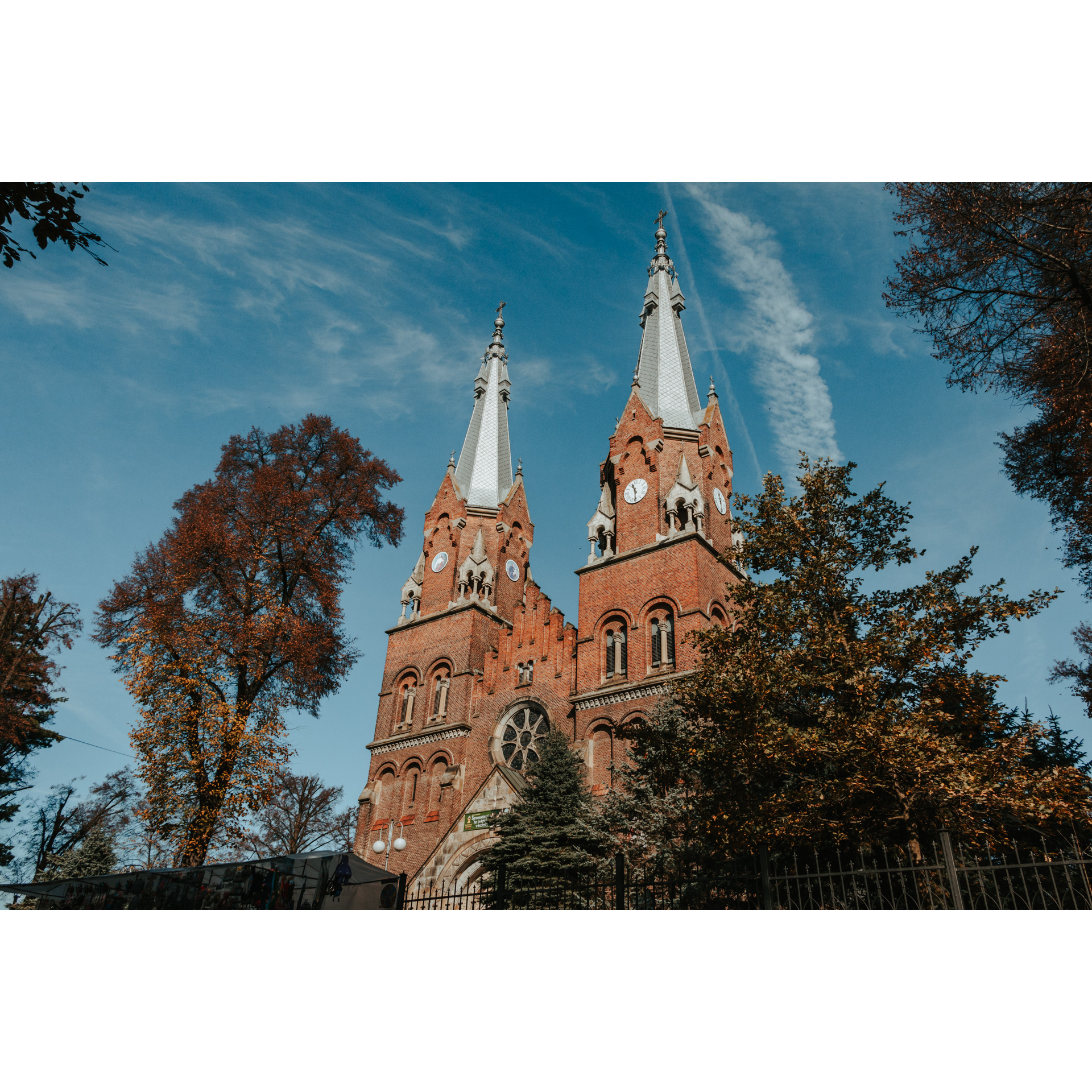 A red brick church with two soaring towers and a round stained-glass window among trees against a blue sky