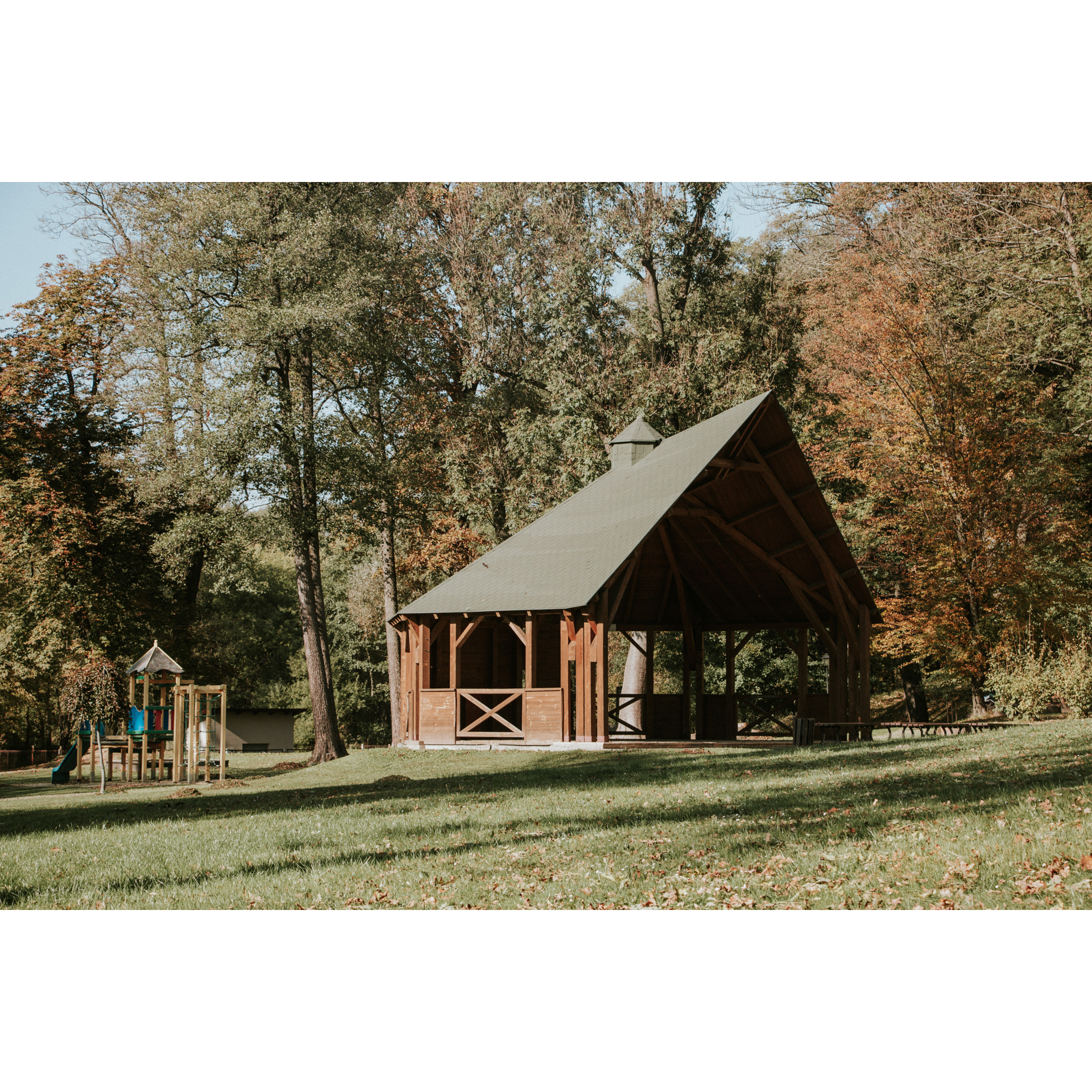 A wooden, open gazebo with a pointed green roof against a background of brown-green trees, a small children's playground on the left