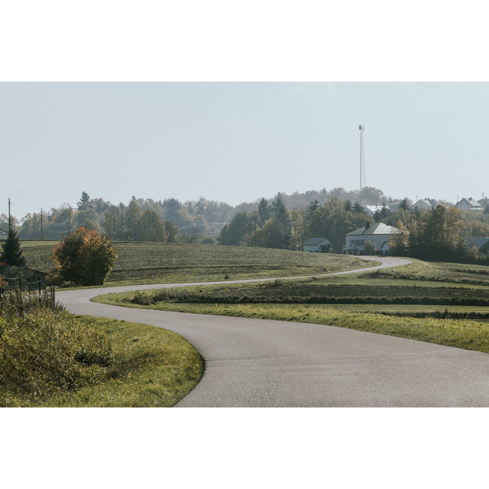 A winding asphalt road running among green meadows, farm buildings, trees and a bright sky in the background