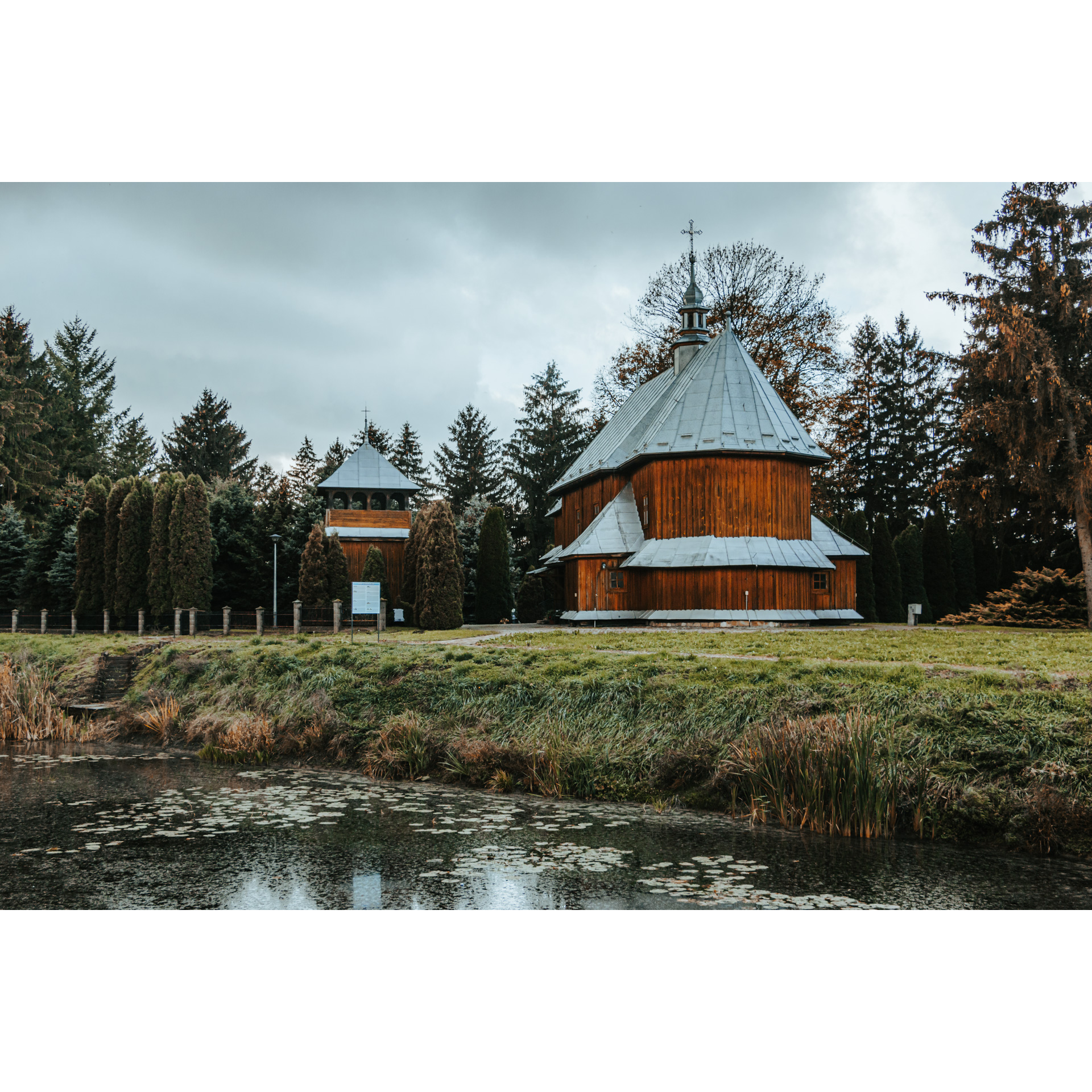 View of the pond and green grass field, in the background a wooden church with a conical, gray sheet metal roof, next to it a belfry with a triangular roof, green trees and bushes around
