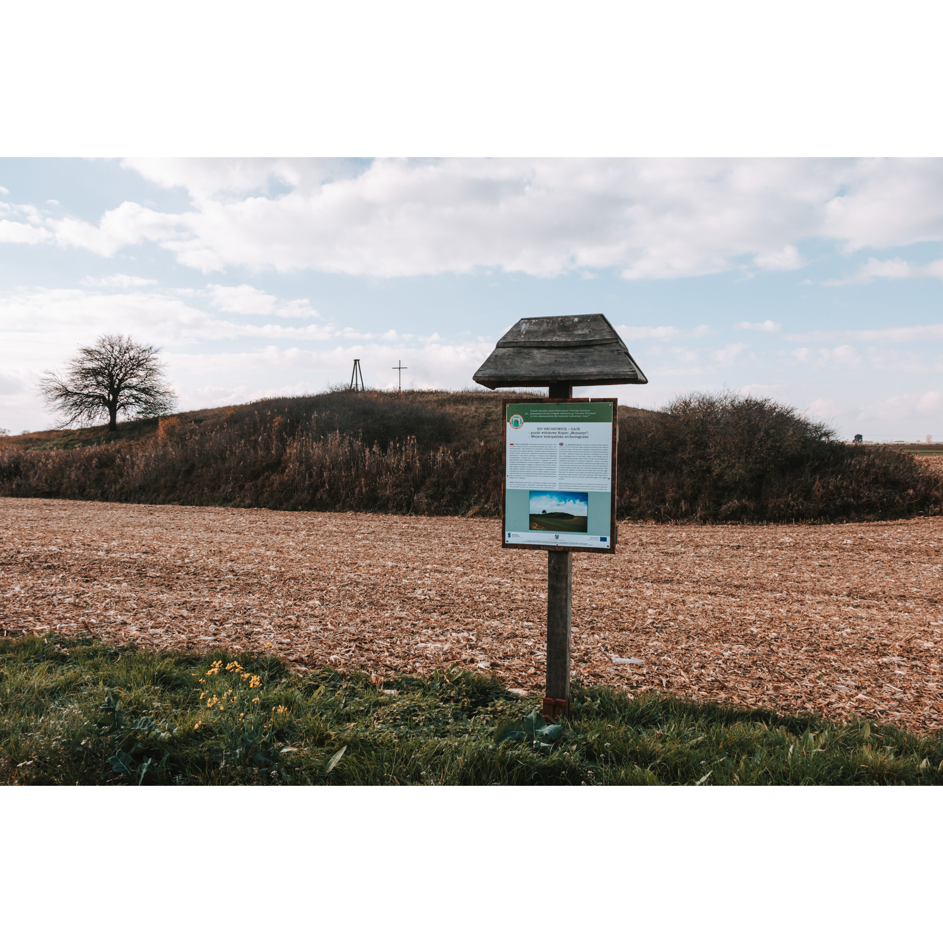 A wooden information board with a roof standing on a plowed field, in the background a hill with a cross and a tree on top, the sky with clouds in the background