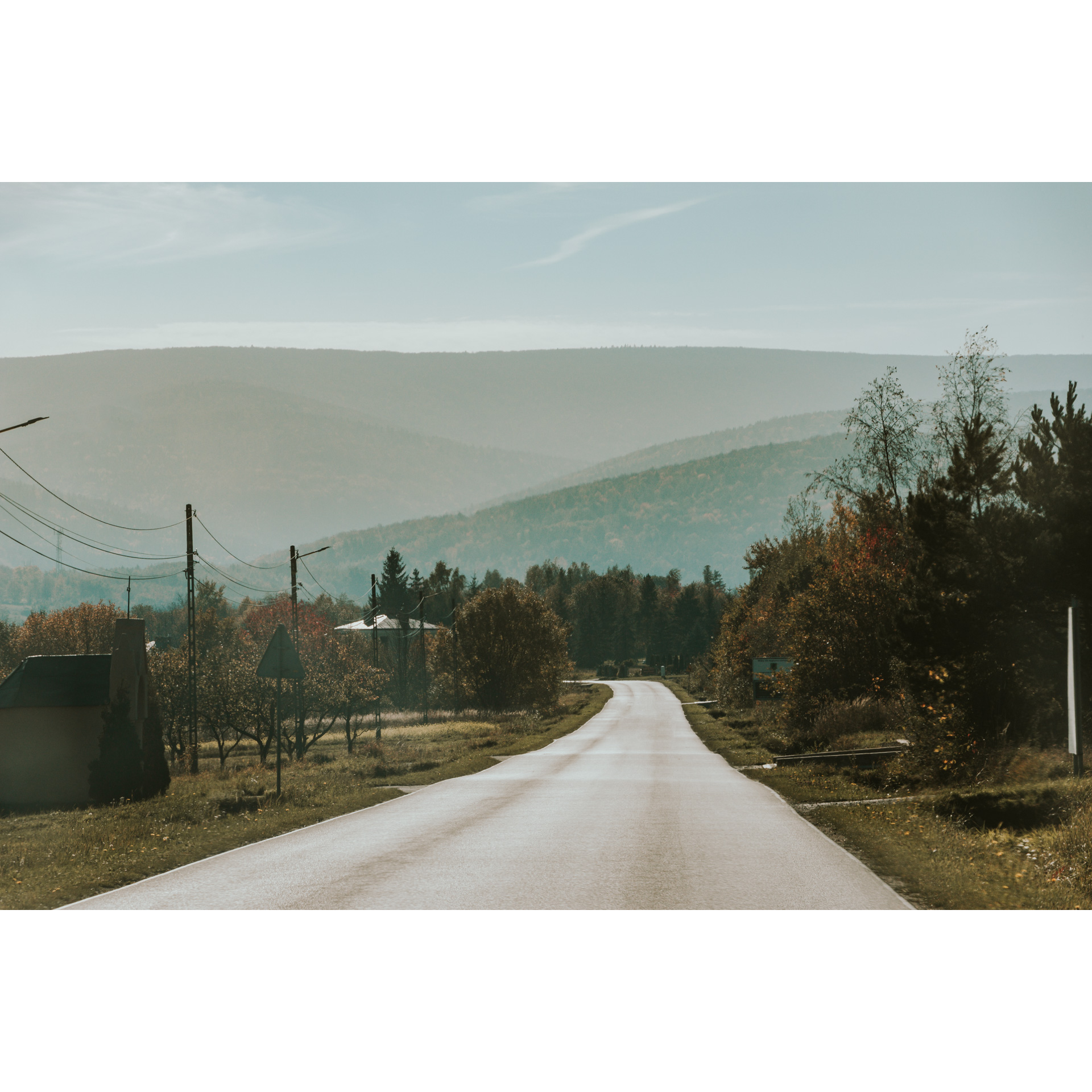 Asphalt road illuminated by the sun, a brick chapel on the roadside, brown-green trees, electric poles, mountains and a bright sky in the distance