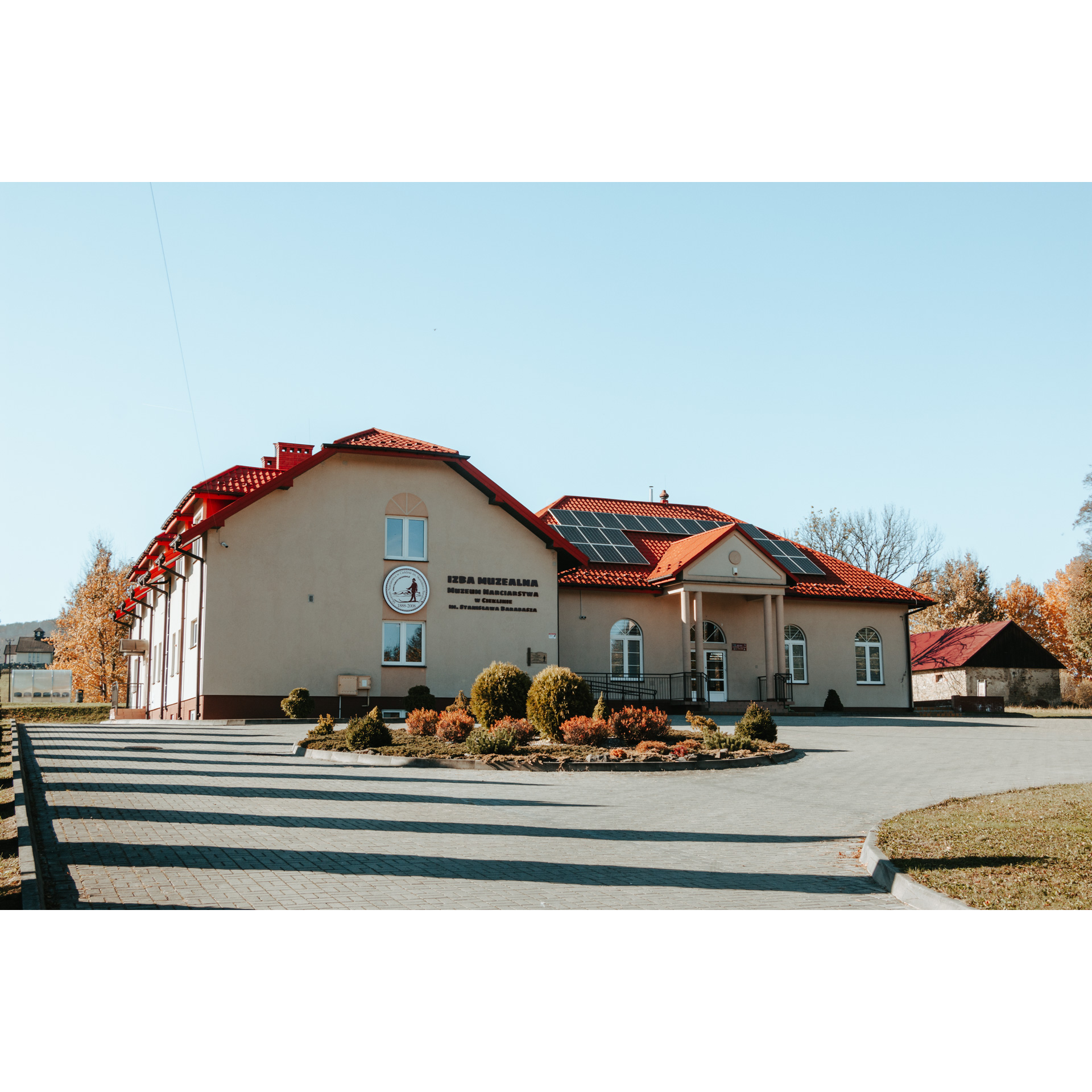 Beige brick buildings with a trapezoidal red roof and columns in front of the entrance, on the side wall a round logo with a man in a hat and skis and the inscription "Izba muzealna"