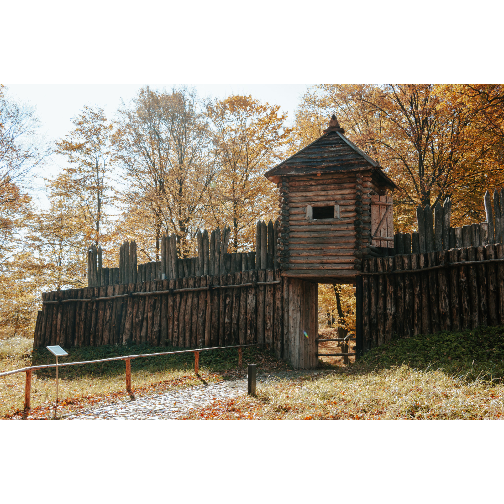 A historic defensive wall built of wooden, pointed logs, an observation box with a roof above the entrance, orange trees in the background