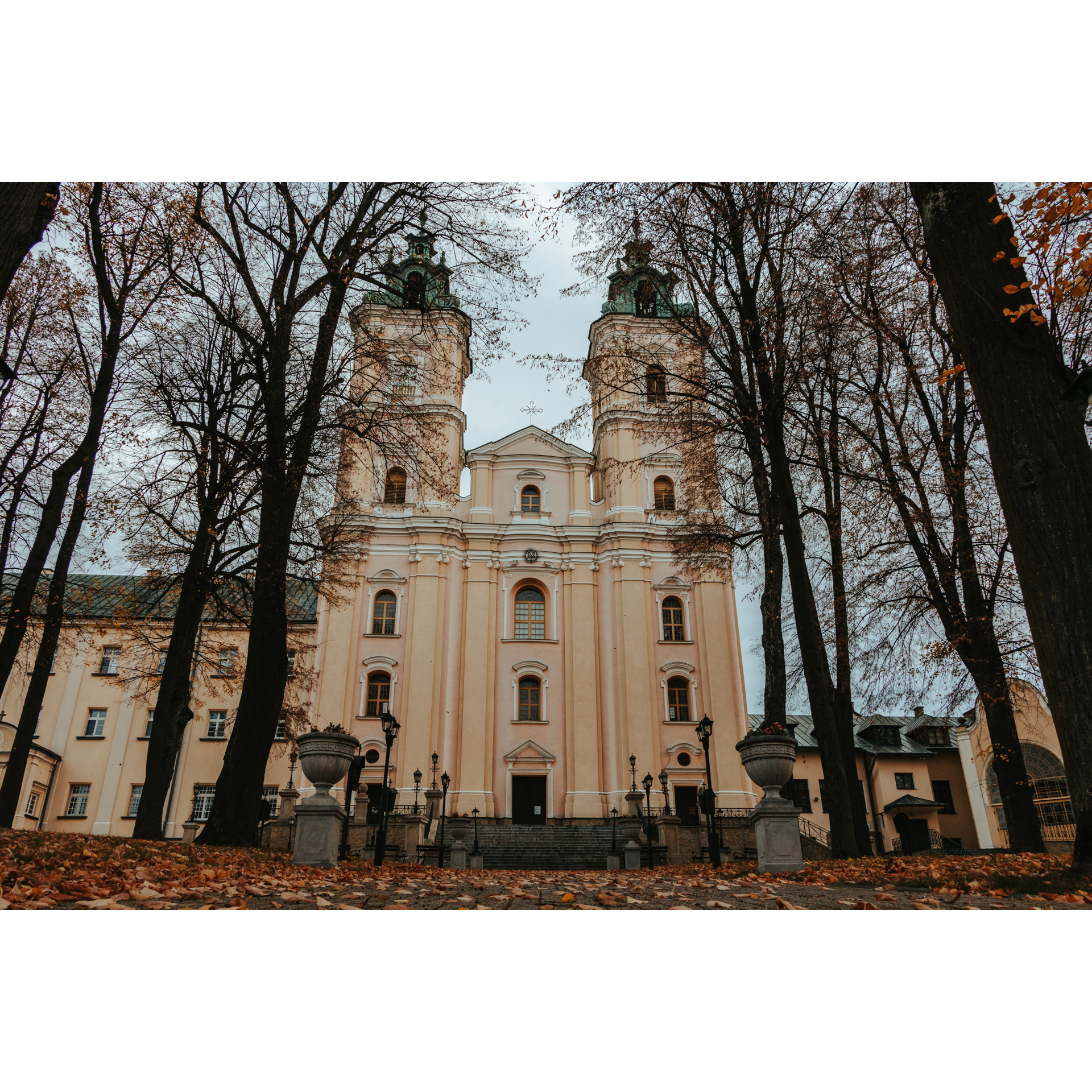 Stojący za drzewami bez liści beżowy, murowany, wysoki kościół z dwoma strzelistymi wieżami zakończonymi krzyżami, fasada z licznymi oknami, po bokach niższe beżowe budynki przylegające do kościoła