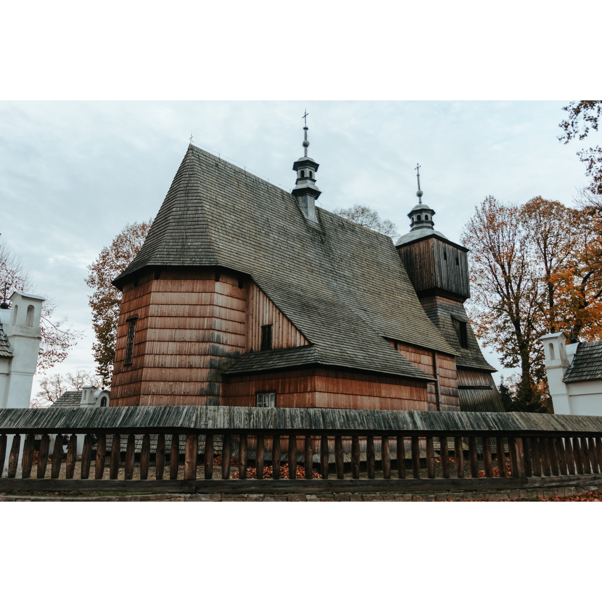 A wooden church with a sloping, dark gray roof and two turrets, a low wooden fence with a roof on the wall in front