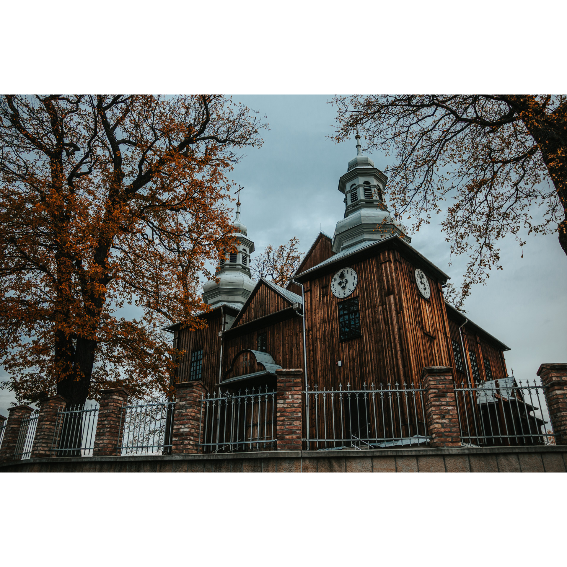 A wooden church with two domed towers with a gray tin roof, a white clock on the facade, tall yellow-brown trees next to it, a metal and brick fence in front of the church