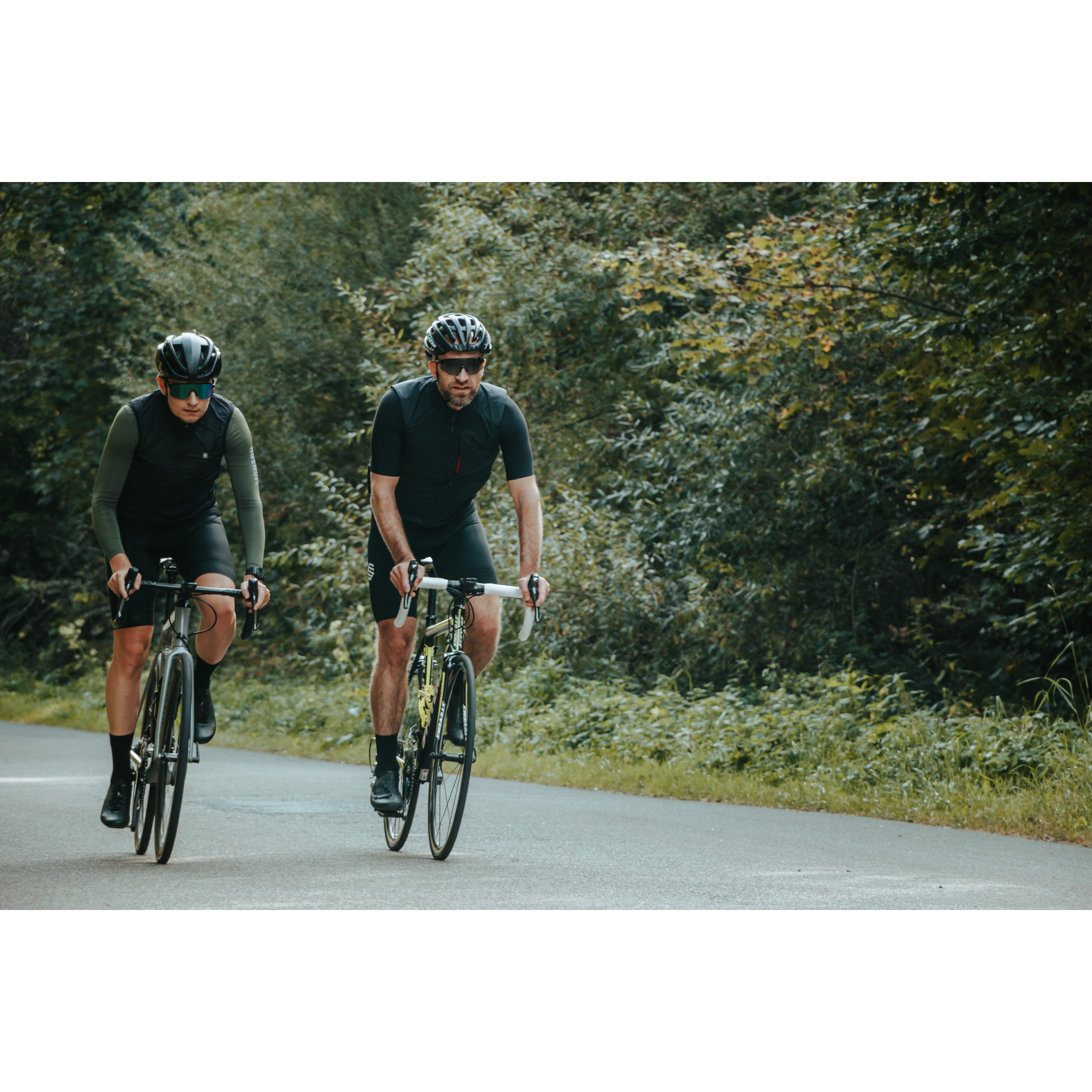 Two cyclists in black cycling clothes, dark glasses and helmets riding bicycles along an asphalt road among green trees and bushes
