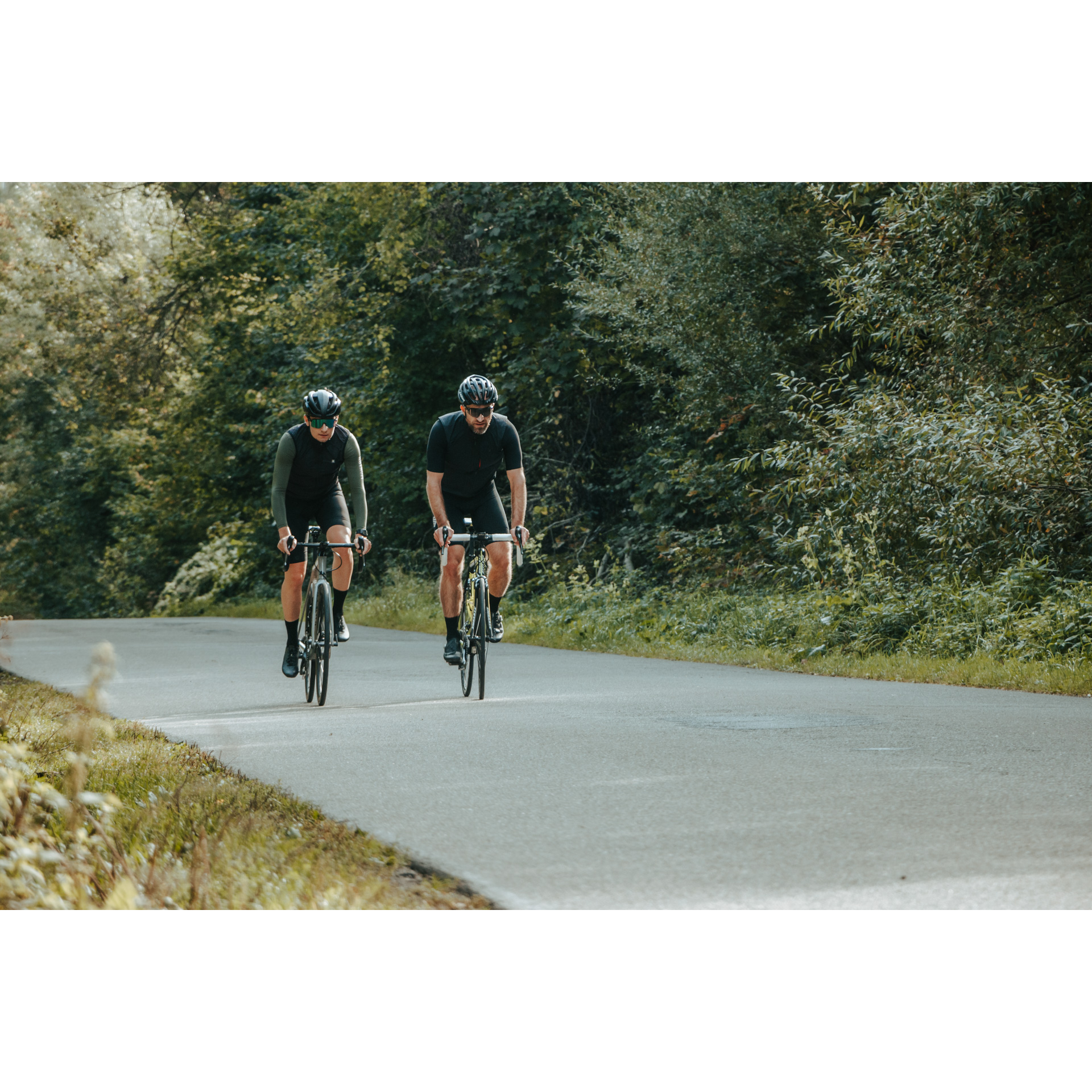 Two cyclists in black cycling clothes, dark glasses and helmets riding bicycles along an asphalt road among green trees and bushes