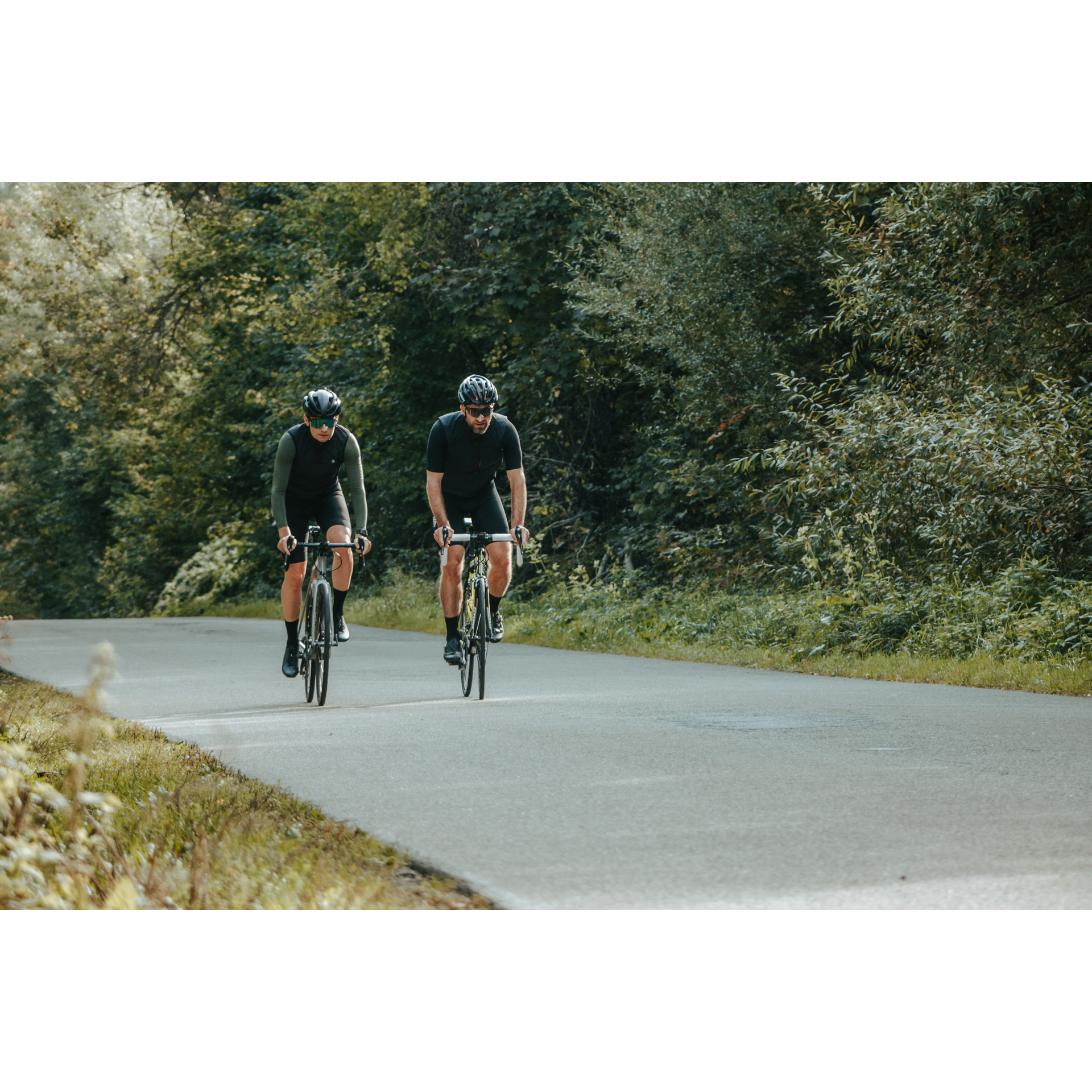 Dwóch rowerzystów w czarnych strojach kolarskich, ciemnych okularach i kaskach jadących na rowerach drogą asfaltową wśród zielonych drzew i krzewów