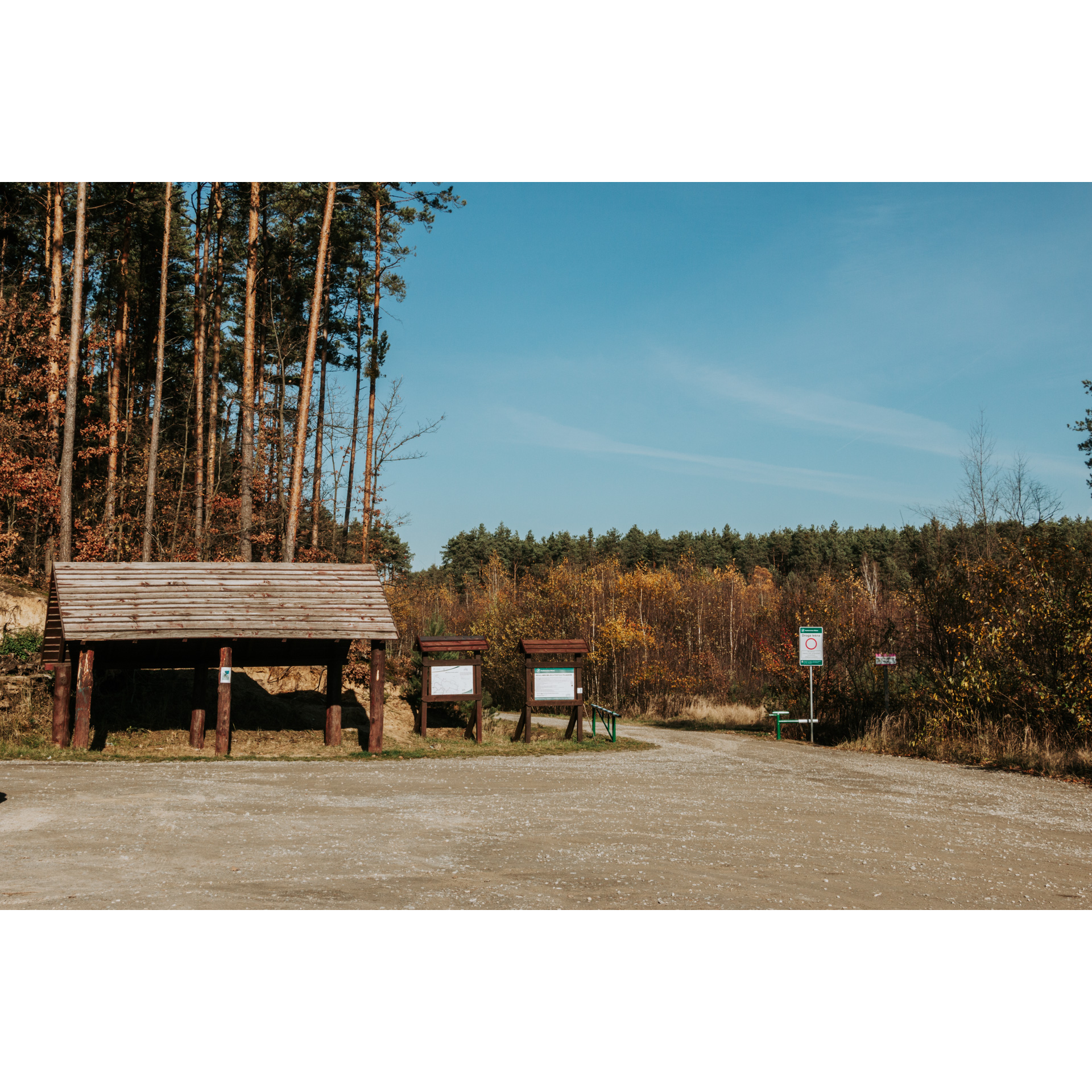 Sandy square with a wooden roof on beams and information boards, tall trees in the background, forest and blue sky in the distance