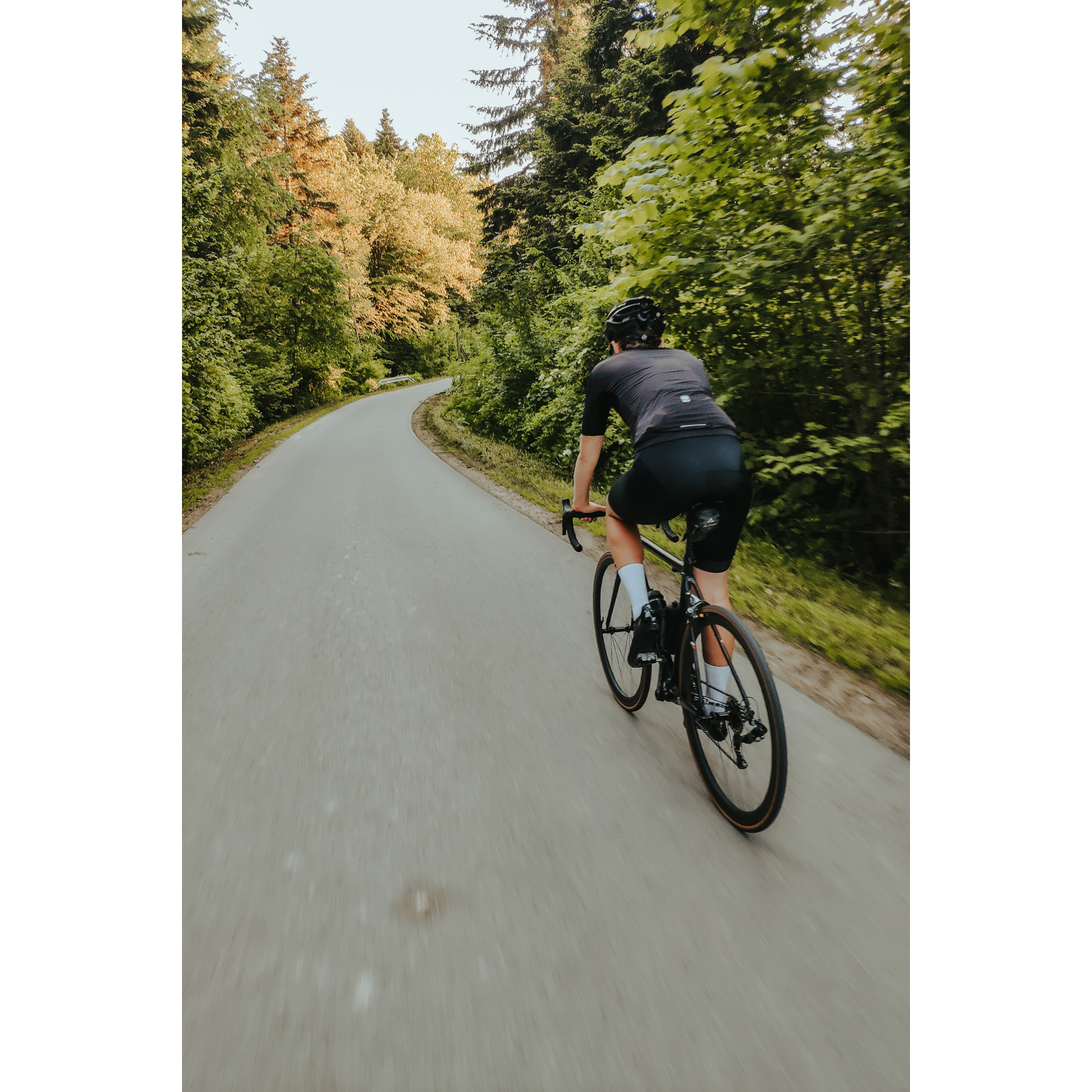 Rowerzysta w czarnym stroju jadący drogą asfaltową przez las pełen drzew z zielonymi liśćmi
