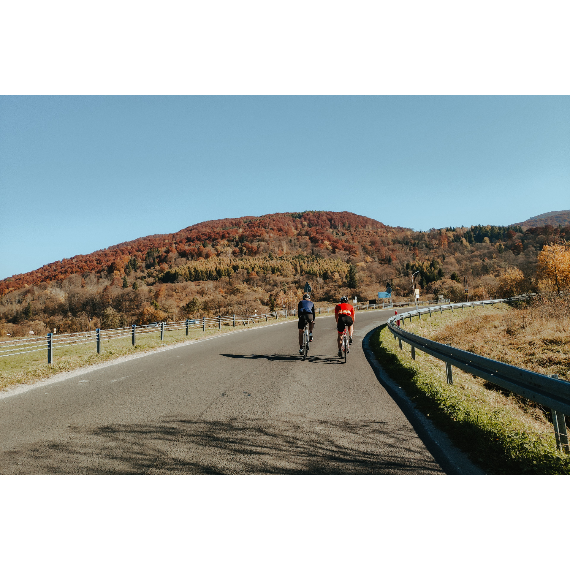 A man dressed in red and a man dressed in blue riding on an asphalt road against the backdrop of a forested mountain with autumn colors