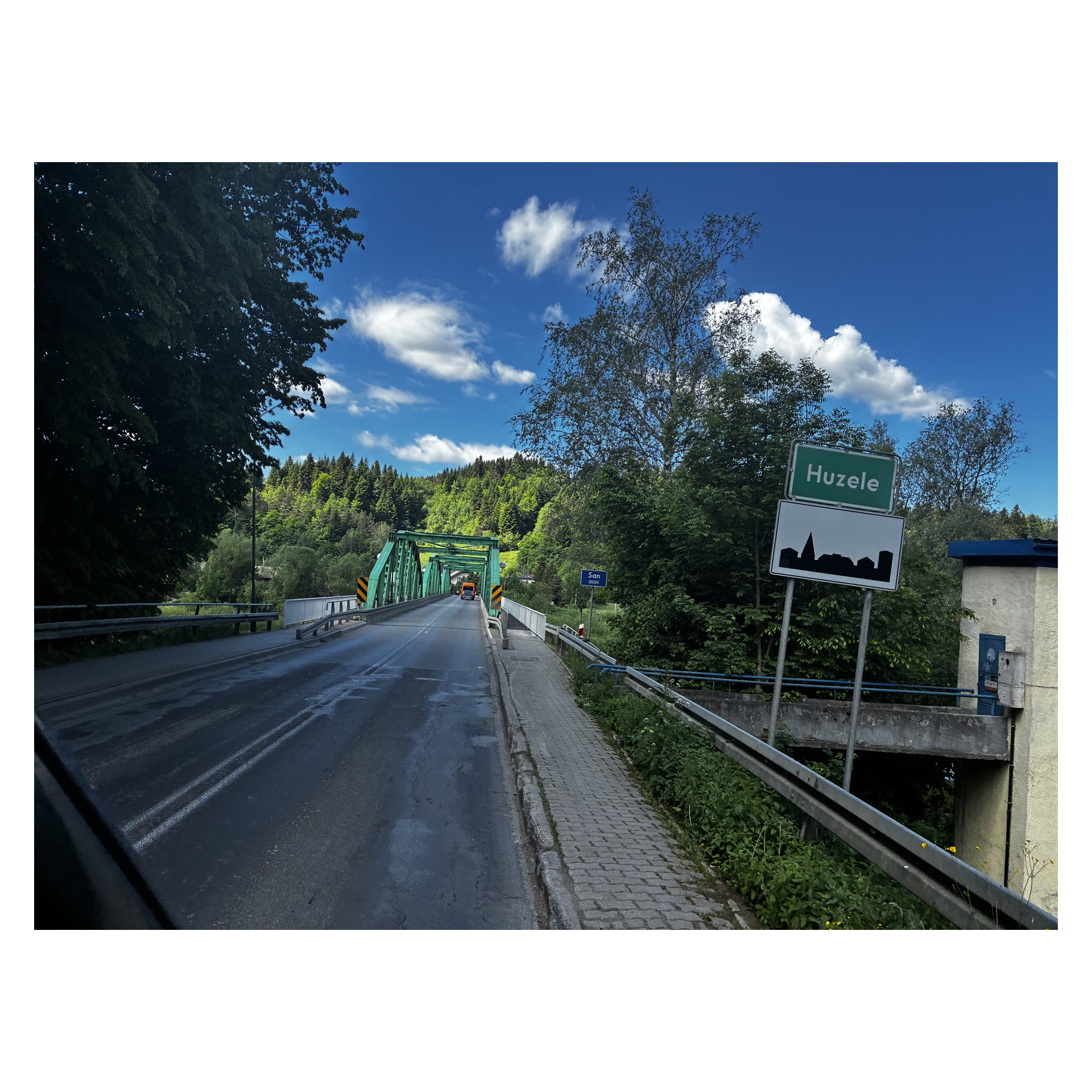 Most na rzece w miejscowości Huzele, zielona stalowa konstrukcja otoczona lasem i niebem z białymi chmurami.