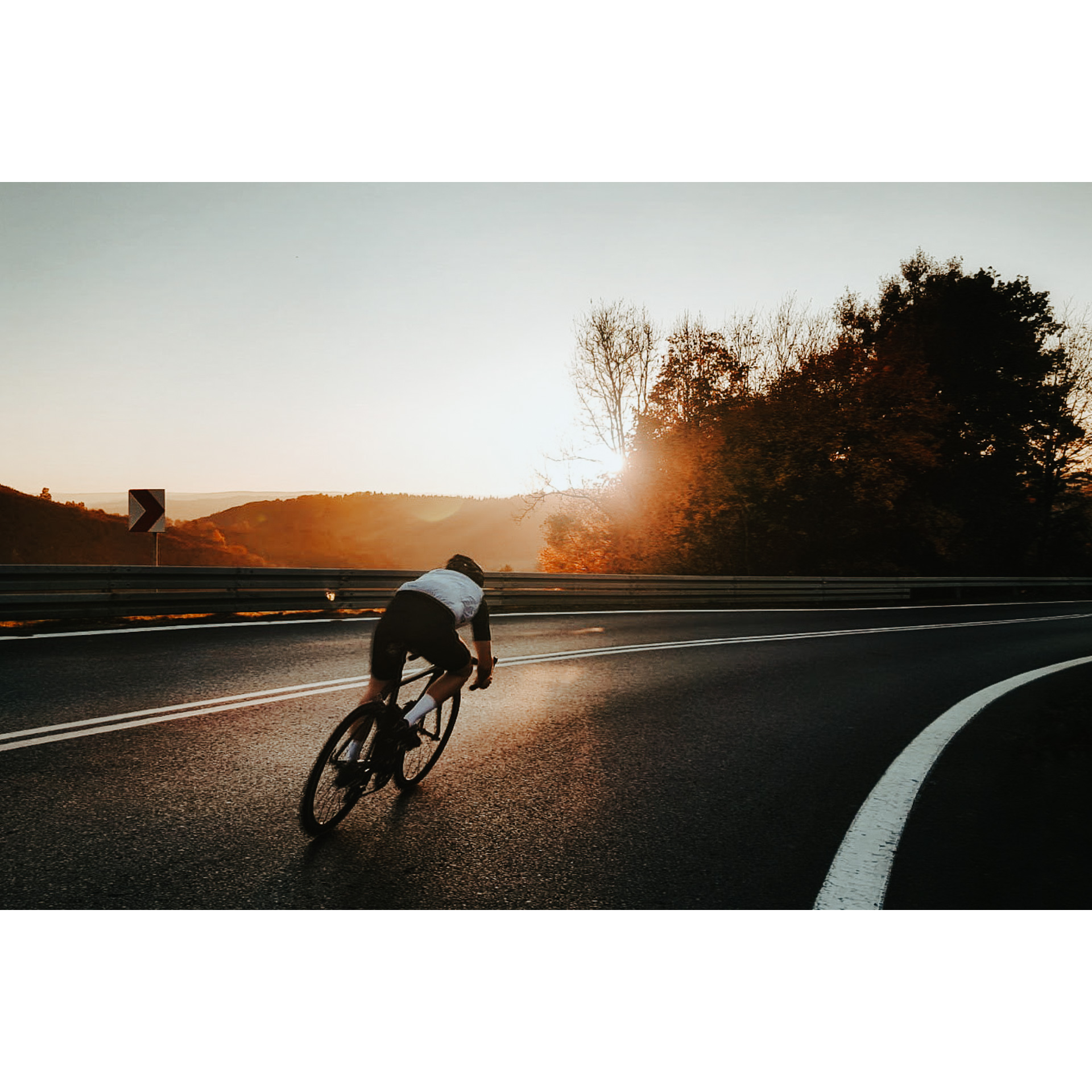 A cyclist in a white T-shirt and black cycling shorts riding on a wide, asphalt road with visible stripes against the setting sun