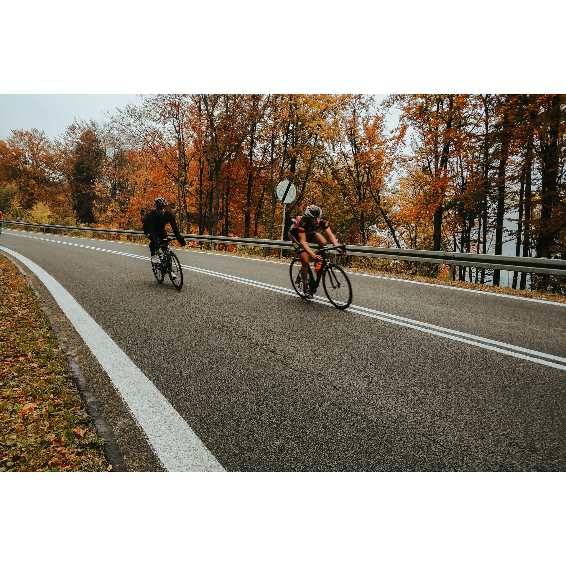 Rowerzyści w czarnych strojach jadący drogą asfaltową w otoczeniu drzew z pomarańczowymi liśćmi 