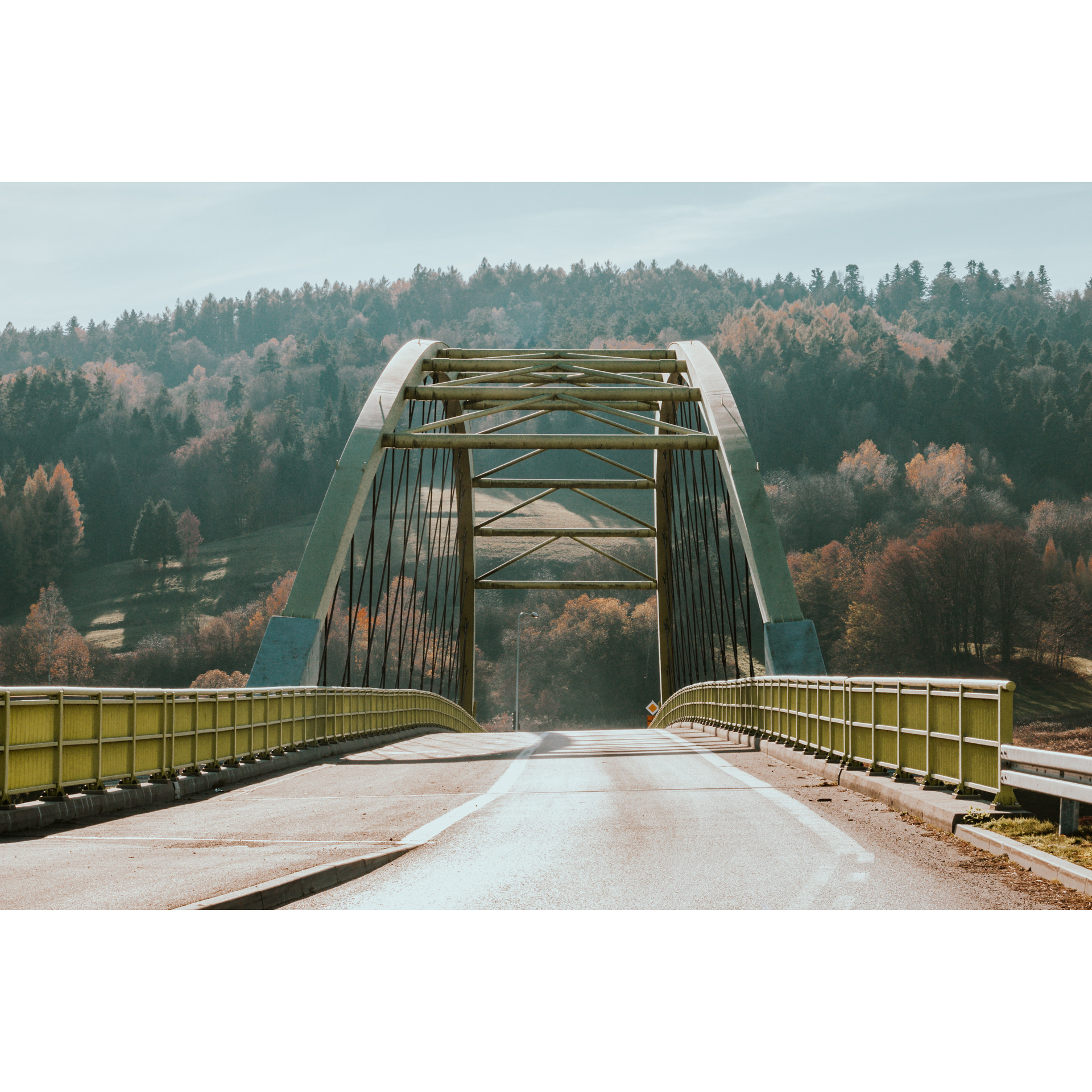 A metal bridge with a semi-circular structure over an asphalt road, trees on a hill in the background