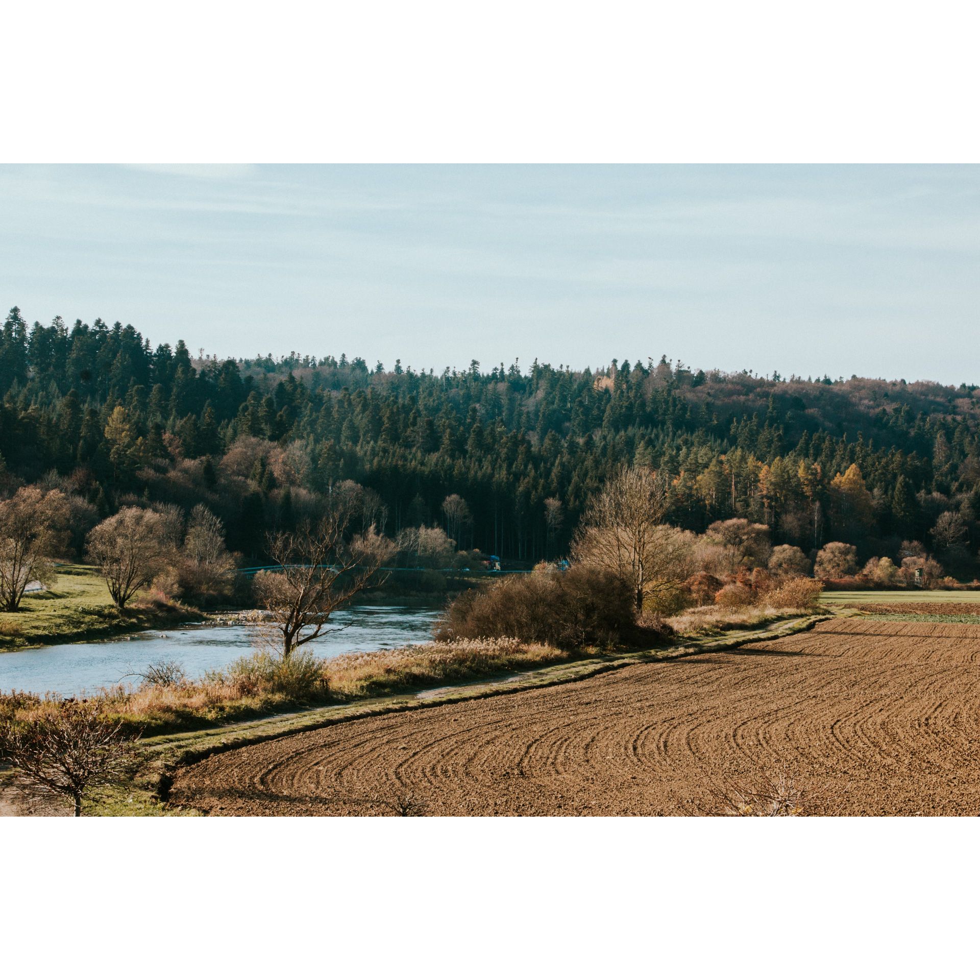 A river flowing along a plowed field, trees on a hill in the background