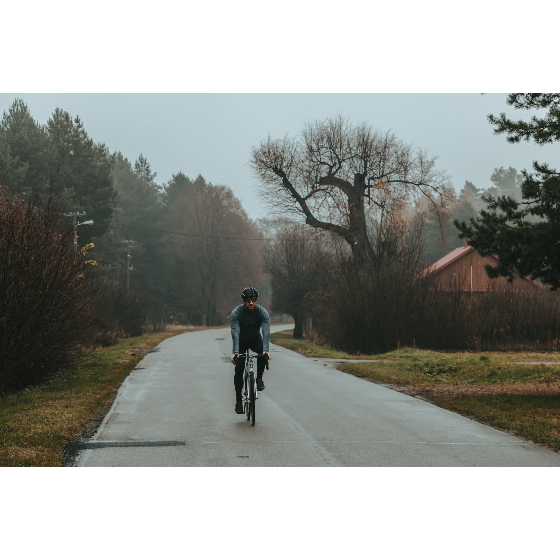 Rowerzysta w niebiesko-czarnym stroju jadący drogą asfaltową w otoczeniu drzew z brązowym, drewnianym budynkiem w tle