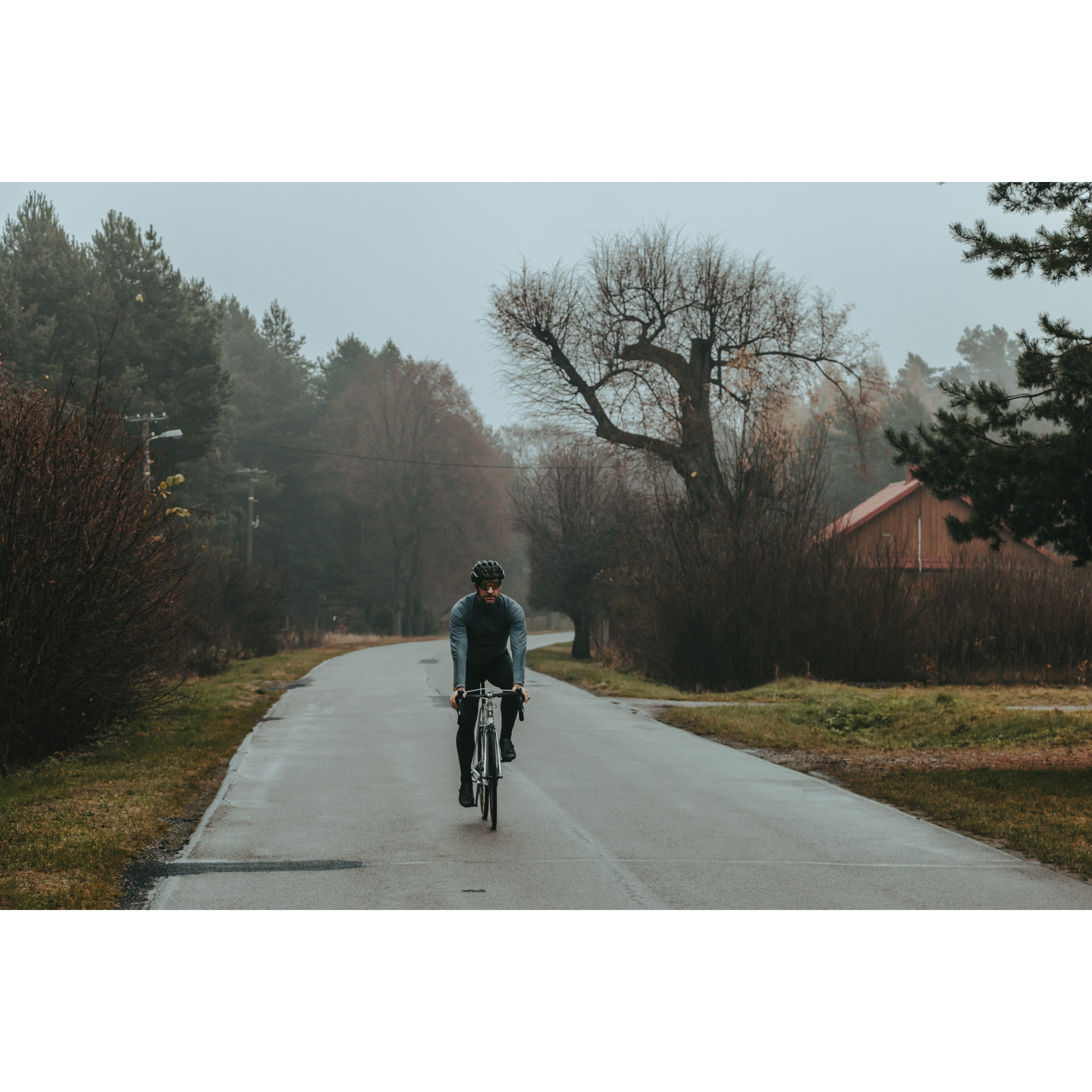 A cyclist in a blue and black outfit riding an asphalt road surrounded by trees with a brown wooden building in the background