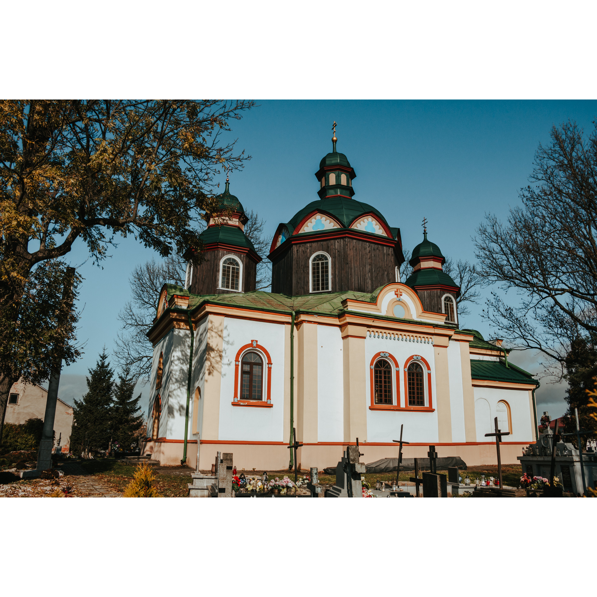 Biała cerkiew z trzema drewnianymi wieżami z zielonym dachem i czerwonymi akcentami wokół okien, dachu i na dole budynku oraz cmentarz na pierwszym planie