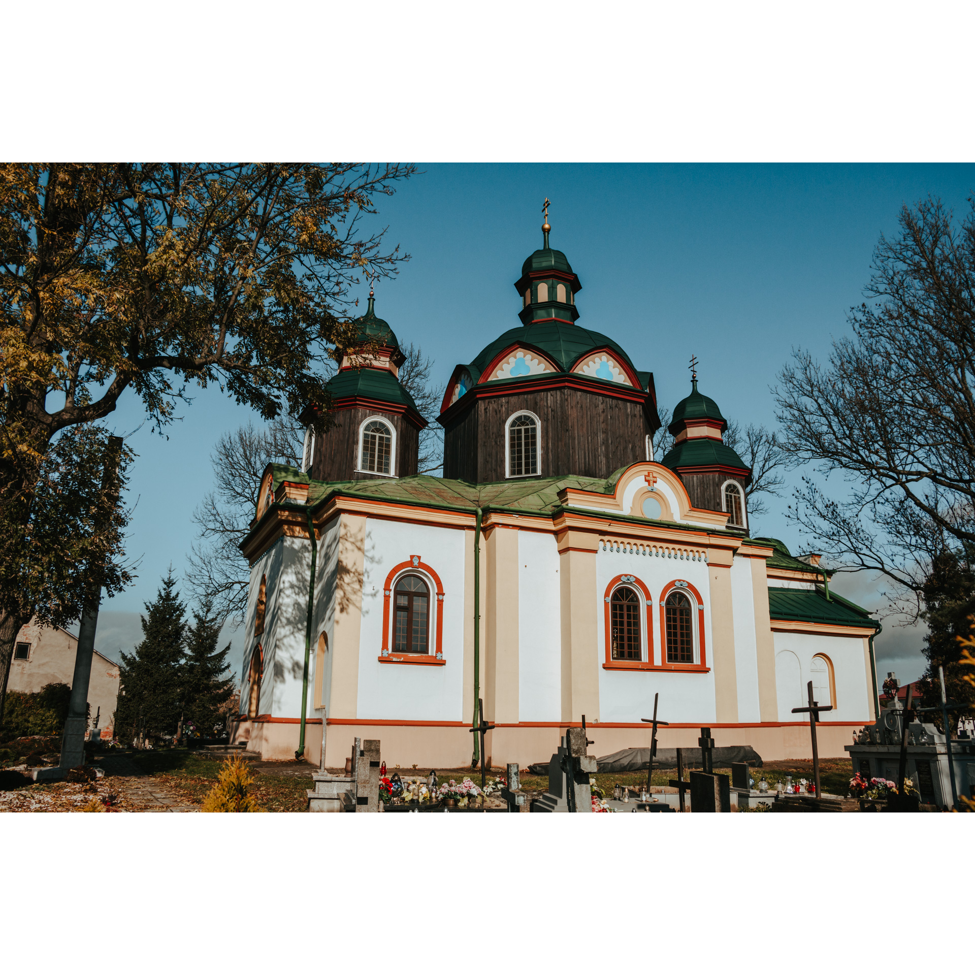 A white orthodox church with three wooden towers with a green roof and red accents around the windows, roof and bottom of the building, and a cemetery in the foreground